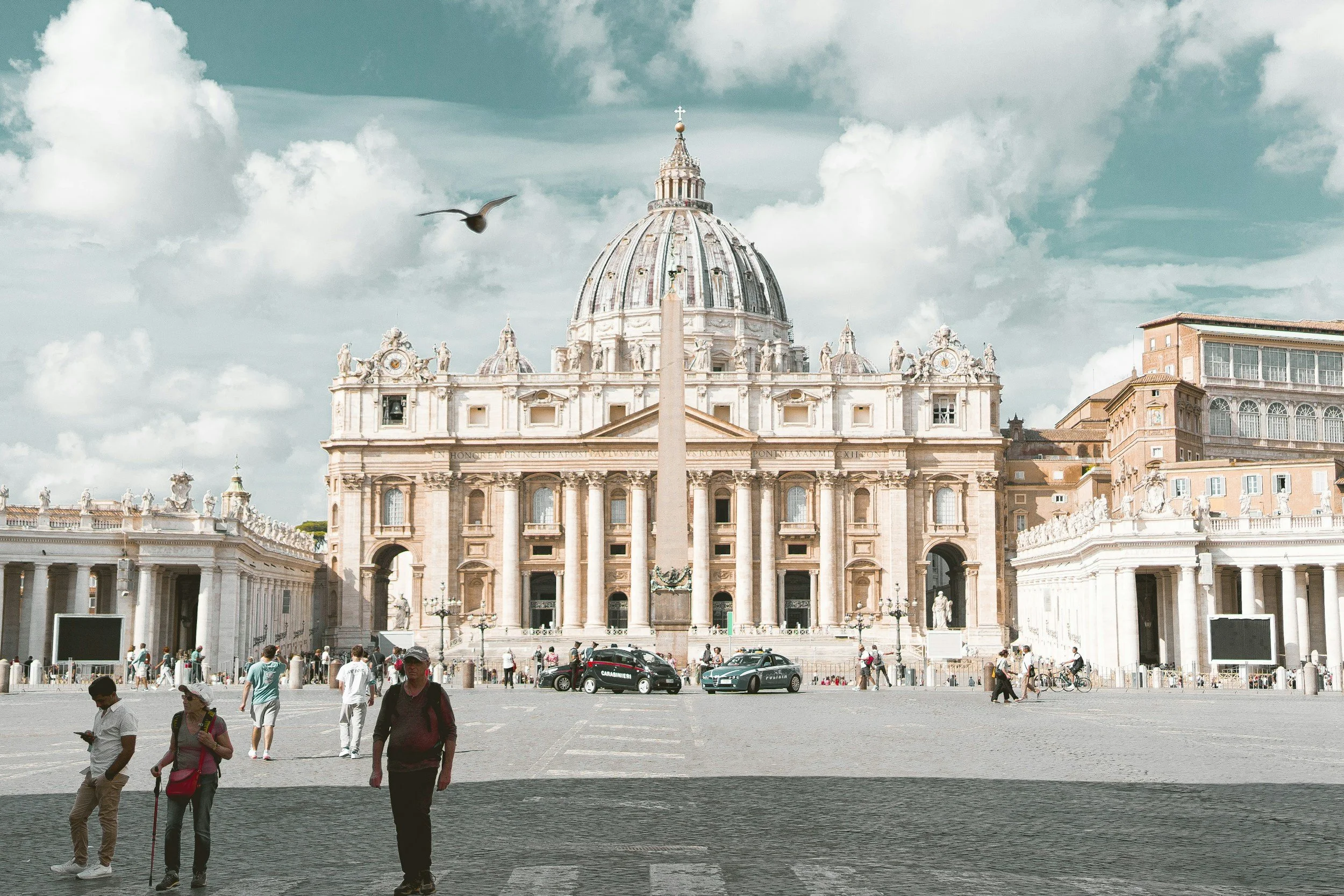 A large historic cathedral with a prominent dome, surrounded by a square with pedestrians, cars, and buildings. The sky is partly cloudy and a bird is flying.