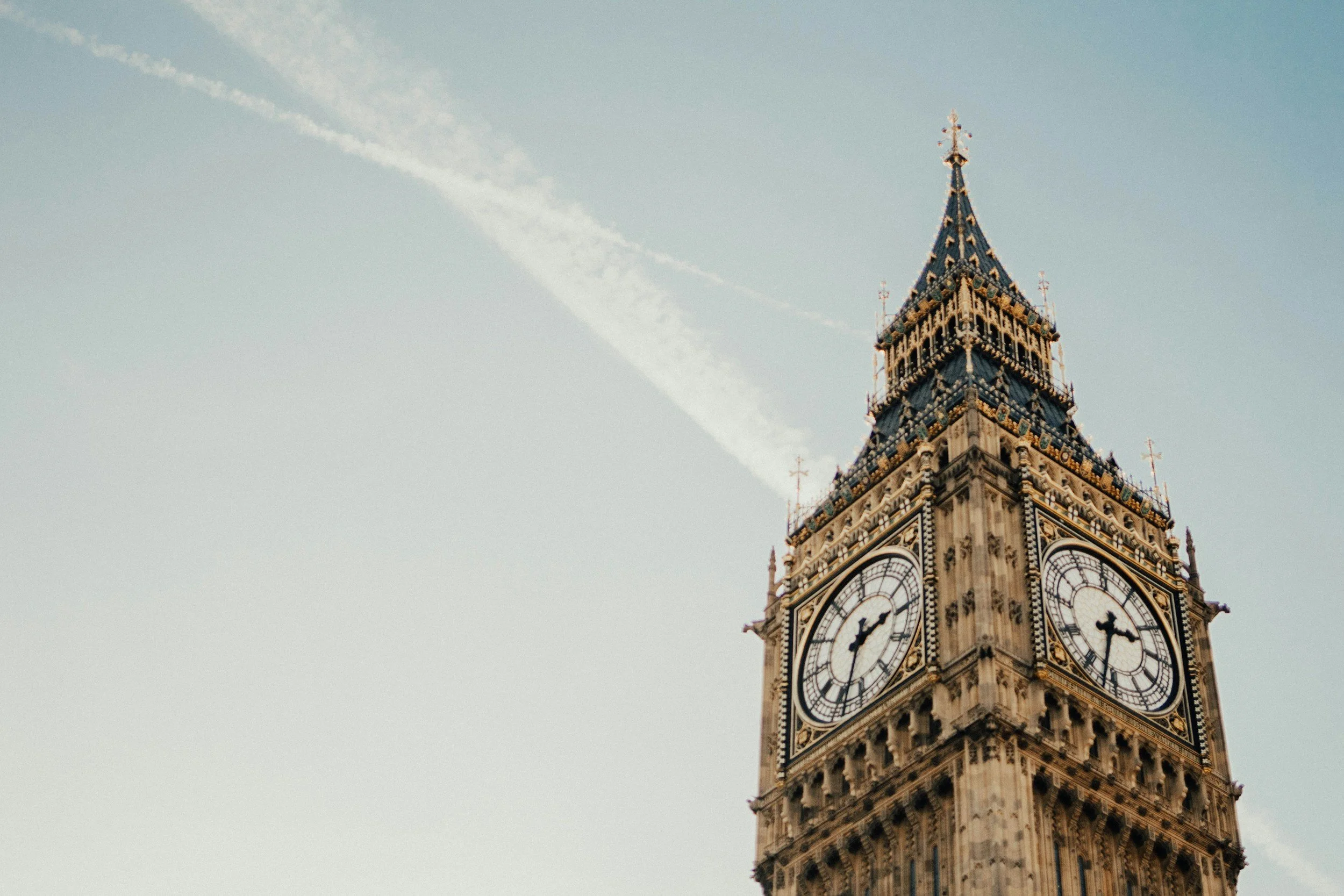 Close-up of Big Ben clock tower in London, showing two clock faces with the time around 4:51, against a light blue sky with wispy clouds and contrails.