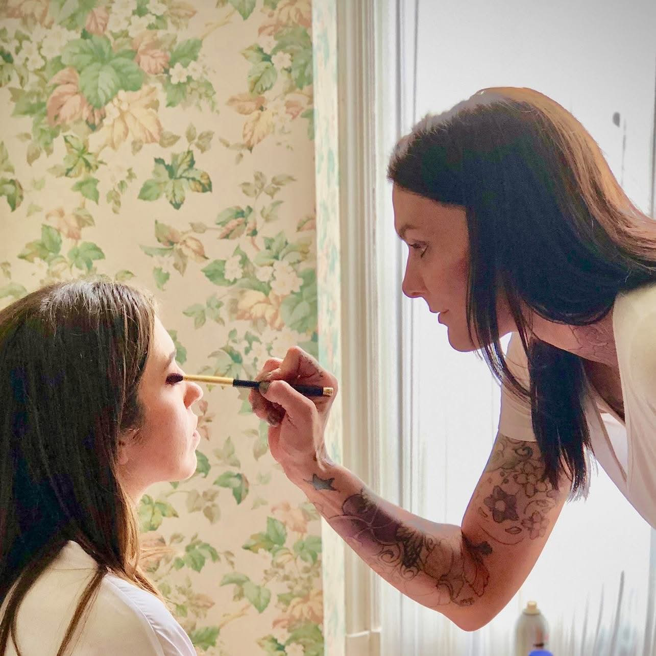 A woman with long dark hair and tattoos on her arm is applying makeup to a young girl with long brown hair sitting in front of her. They are in a room with floral wallpaper and light from a nearby window.