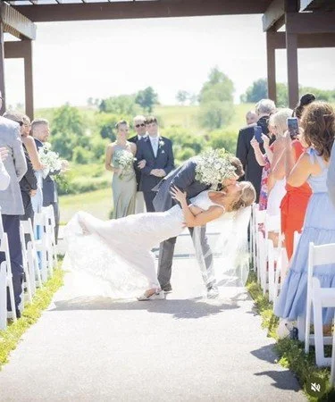 A bride and groom sharing a kiss at their outdoor wedding ceremony, with guests capturing the moment on their phones.