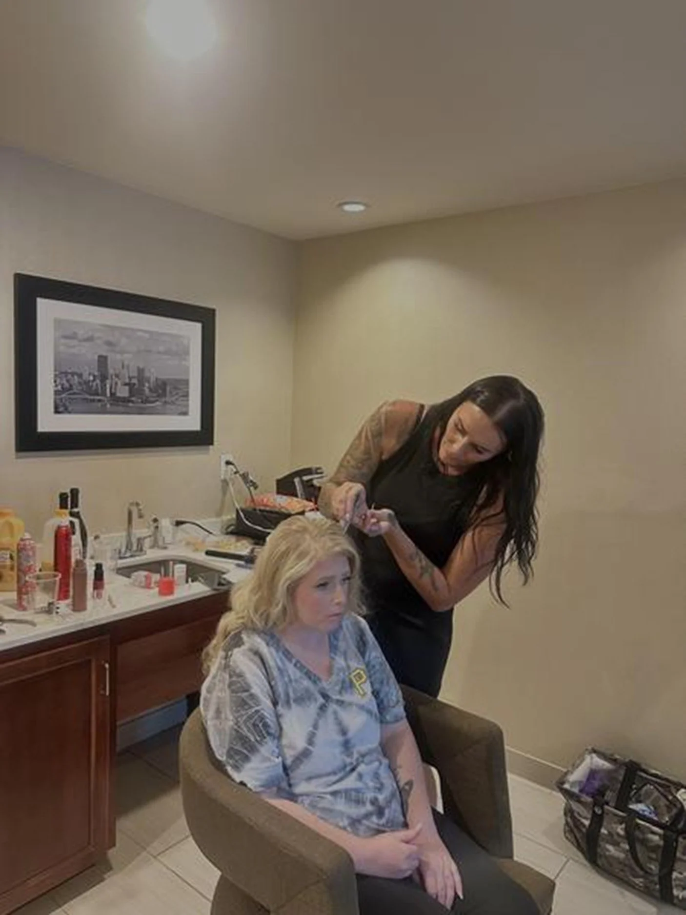A woman with long dark hair and tattoos is applying makeup or styling to a blonde woman sitting in a chair in a room with beige walls and a black and white cityscape picture on the wall.