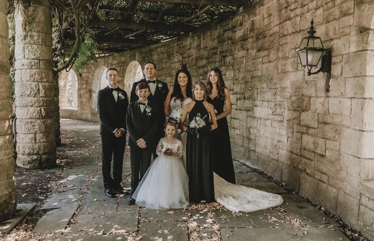 Group of six women, including a bride in a white gown, and five bridesmaids or family members in pink dresses, standing in front of a wooden barn door. They are holding bouquets and smiling.