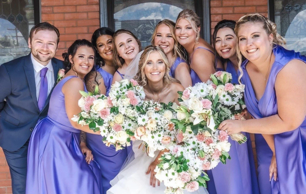 A bride and five bridesmaids standing outdoors on a grassy field, smiling and holding bouquets of white flowers.