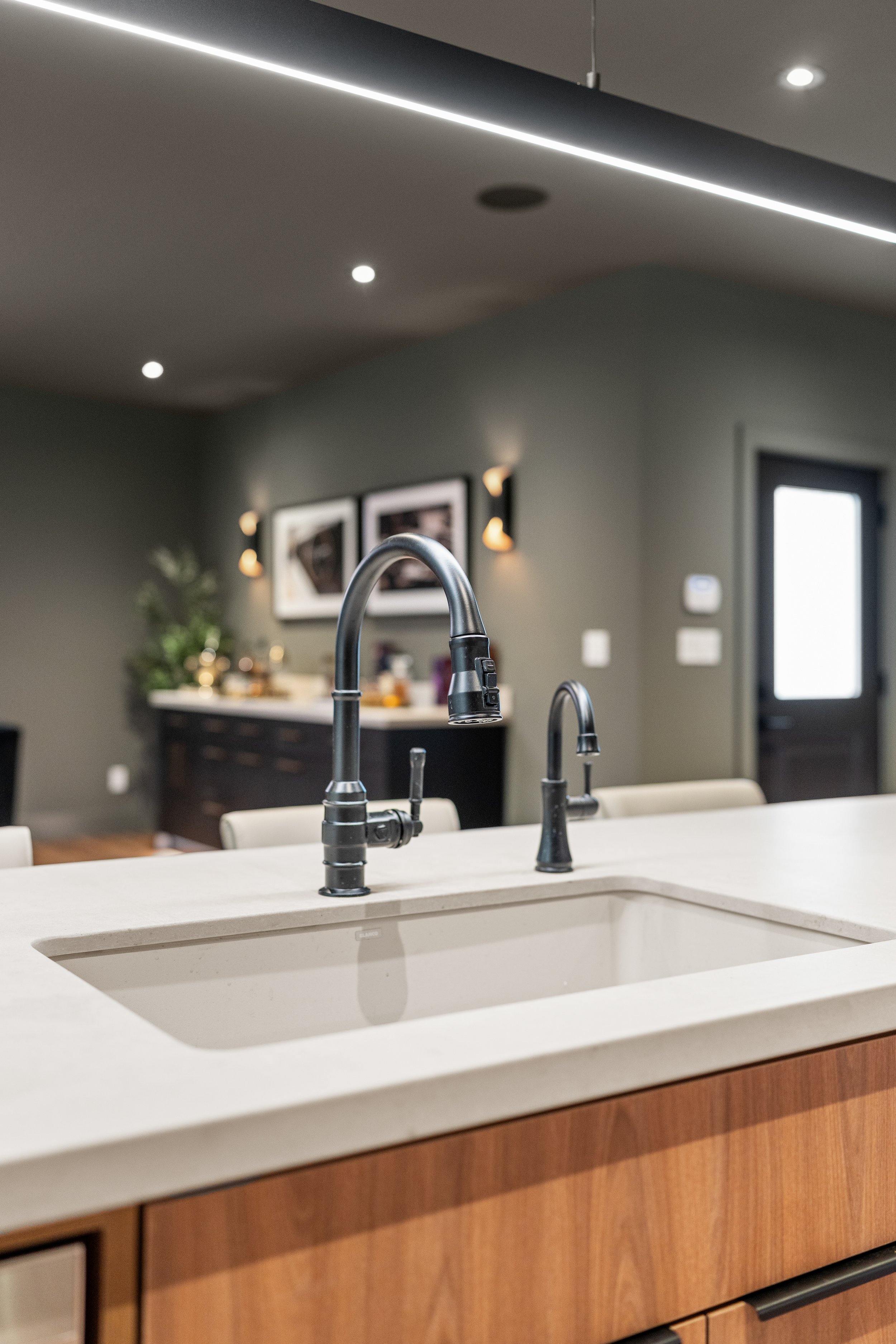 Modern kitchen with black faucet, white countertop, wooden cabinetry, framed pictures on green wall, and entry door in background.