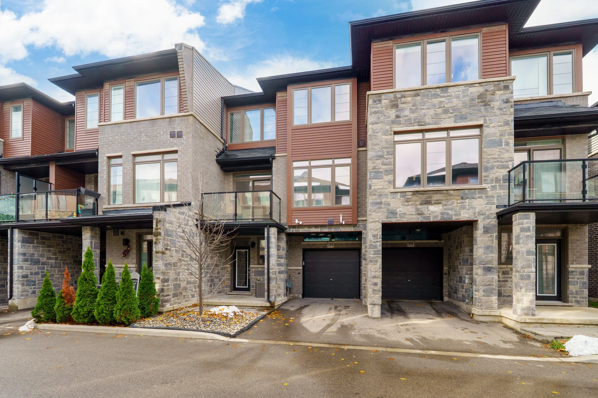 Multi-story modern apartment building with stone and wood siding, glass balconies, and black garage doors under a partly cloudy sky.