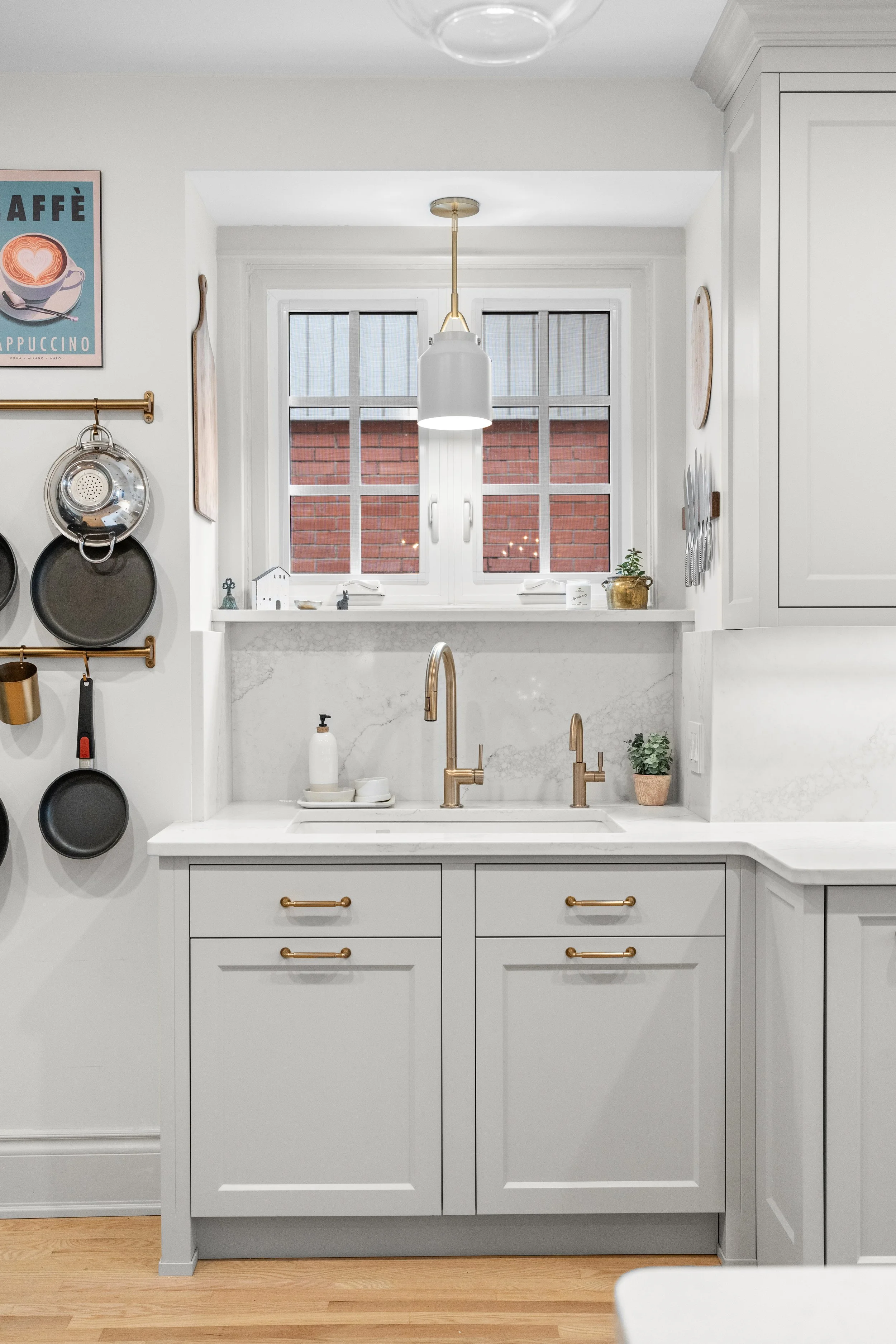 Modern kitchen with light gray cabinets, gold hardware, white marble countertops, a stainless steel double sink with gold faucet, and a window above. To the left, hooks hold black pans and a stainless steel colander. There are small potted plants and