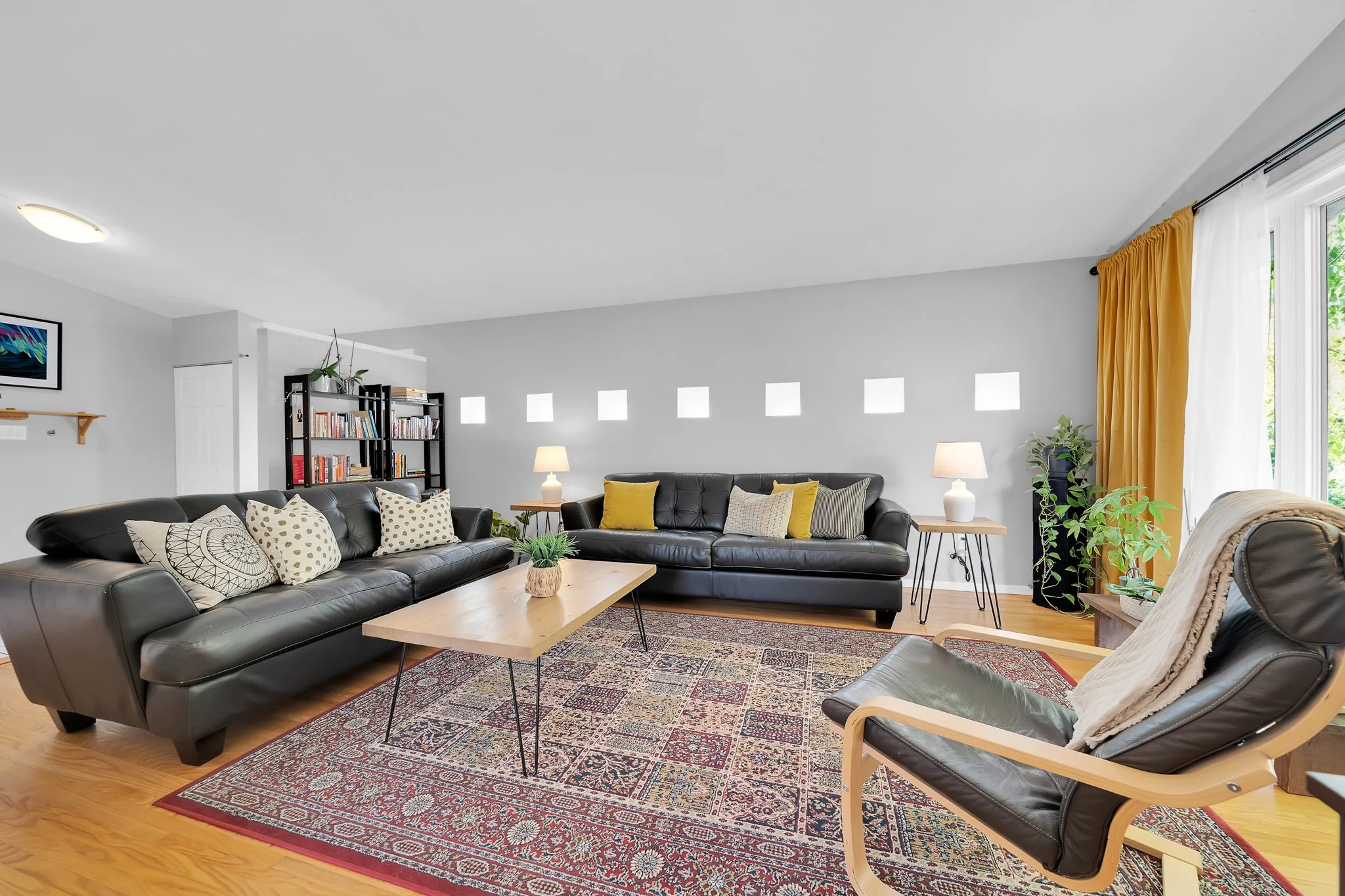 Living room with three black leather sofas, a wooden coffee table, patterned area rug, and yellow curtains. Decor includes pillows, lamps, plants, and a bookshelf.