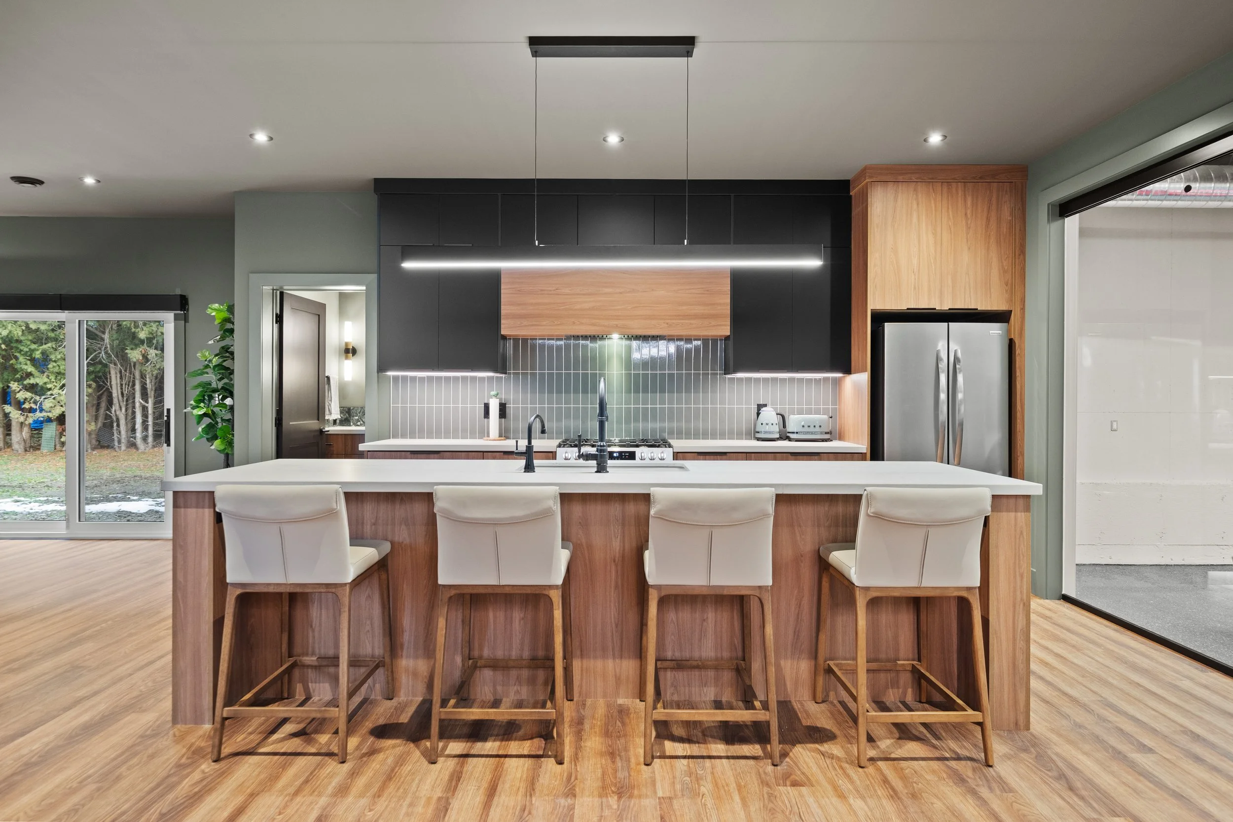 Modern kitchen with white island, four beige barstools, black and wooden cabinets, gray tiled backsplash, stainless steel appliances, and sliding glass doors leading outside.