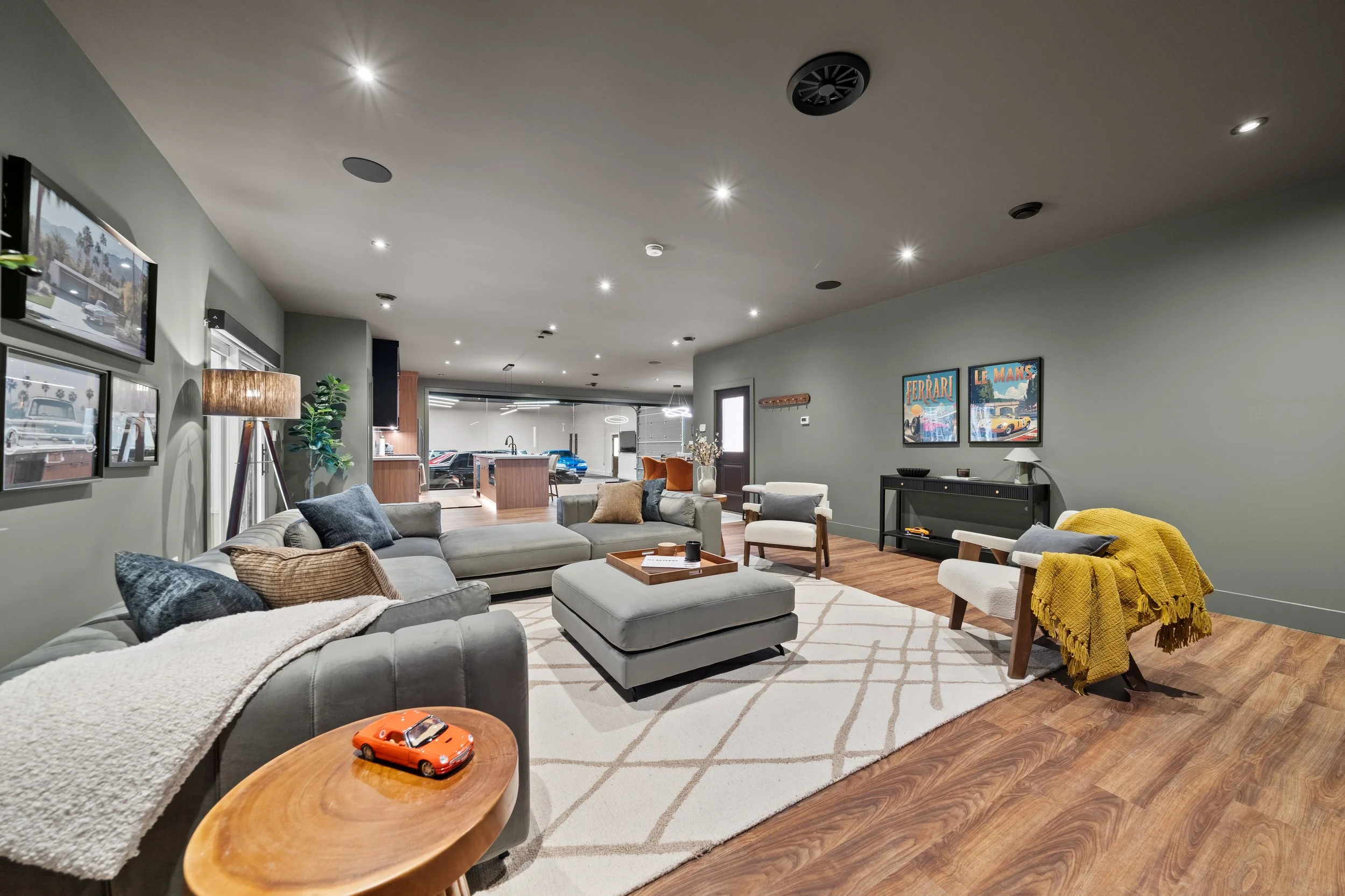 Modern living room with gray sectional sofa, white armchair, area rug, framed car posters, and a view into an open garage showing a blue sports car.