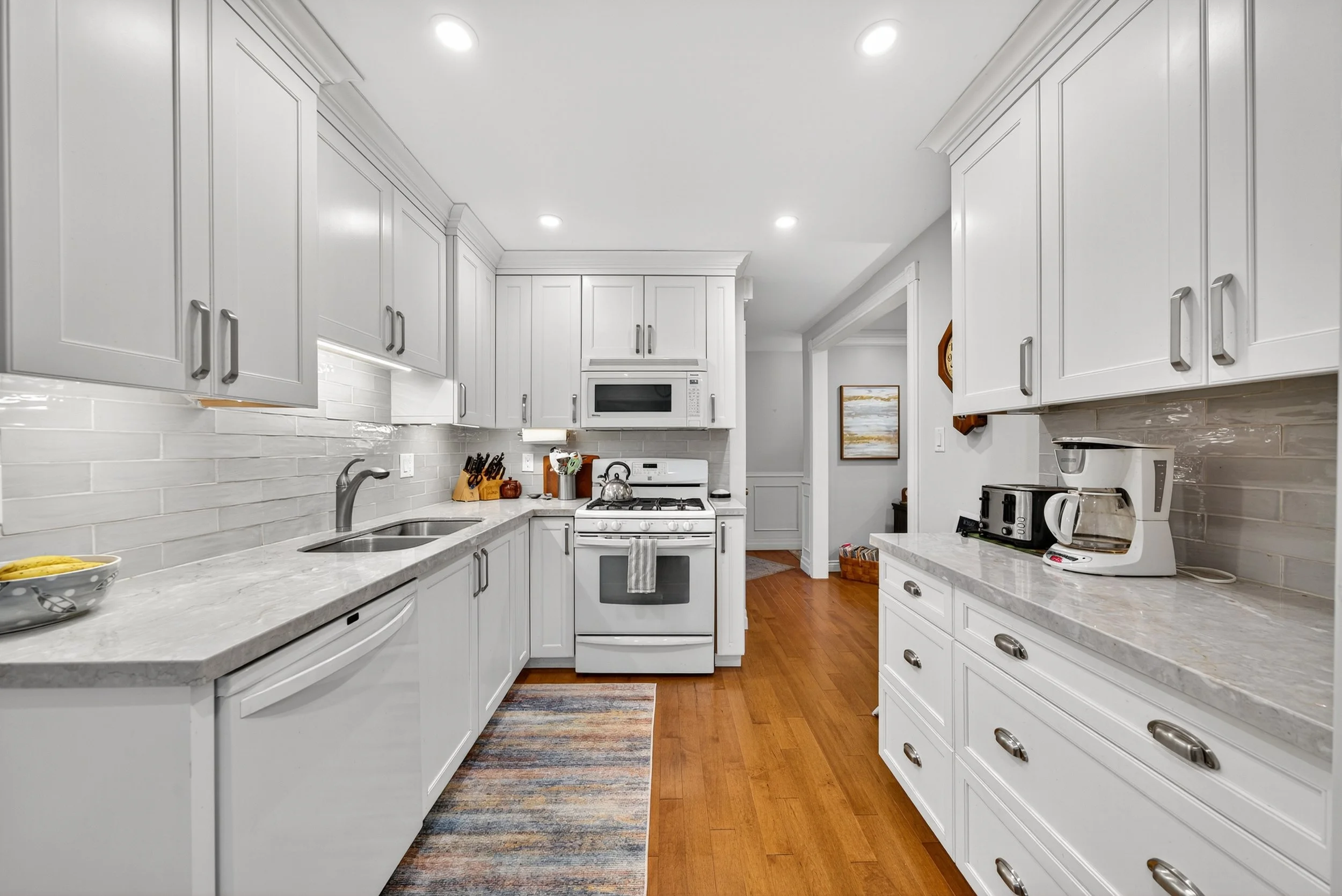 Bright kitchen with white cabinets, marble countertops, and stainless steel appliances including microwave, stove, toaster, and coffee maker, with hardwood floors and a colorful rug.
