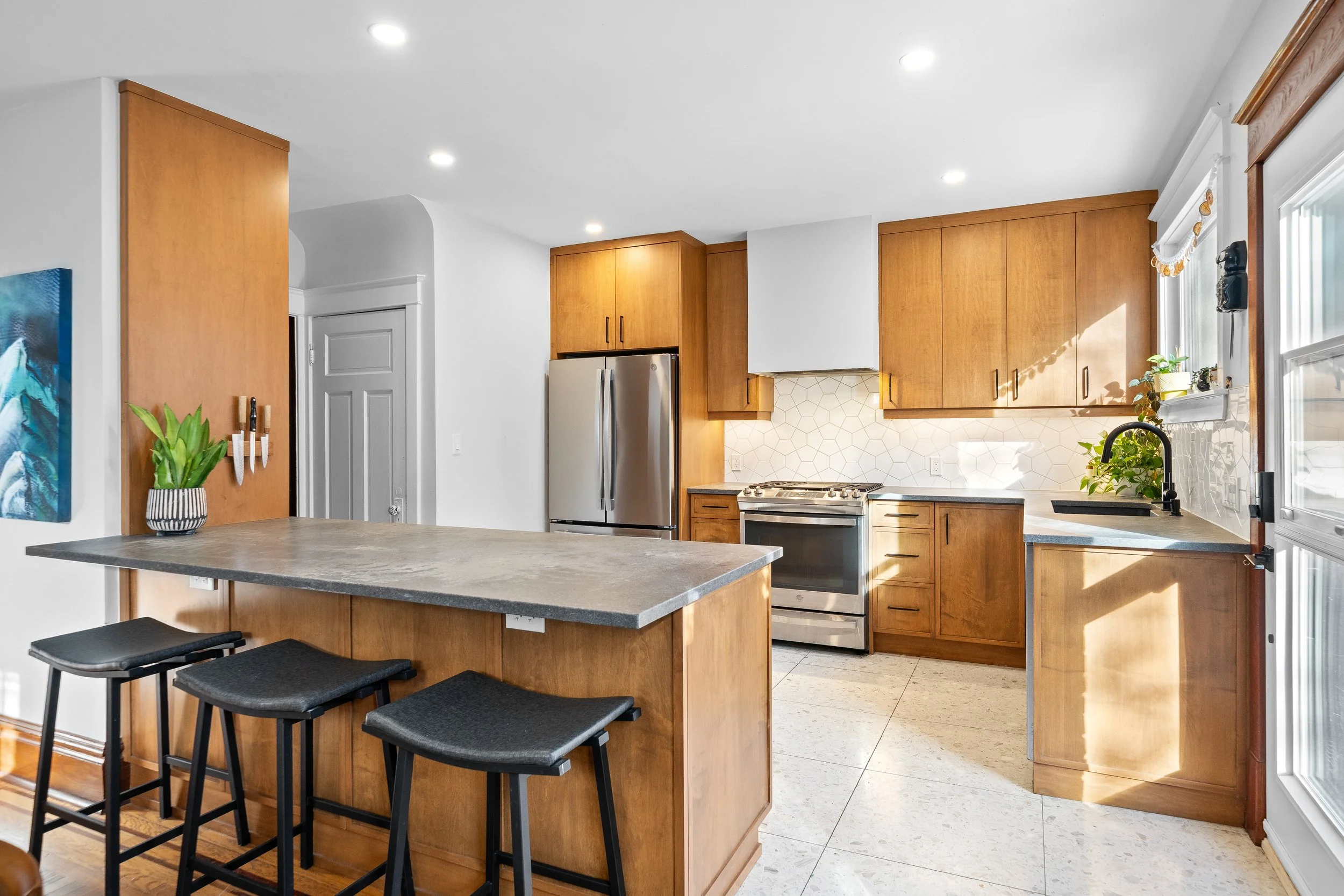 Kitchen with wooden cabinets, stainless steel refrigerator, stove, black countertops, tiled backsplash, window with plants, and a kitchen island with three black stools.