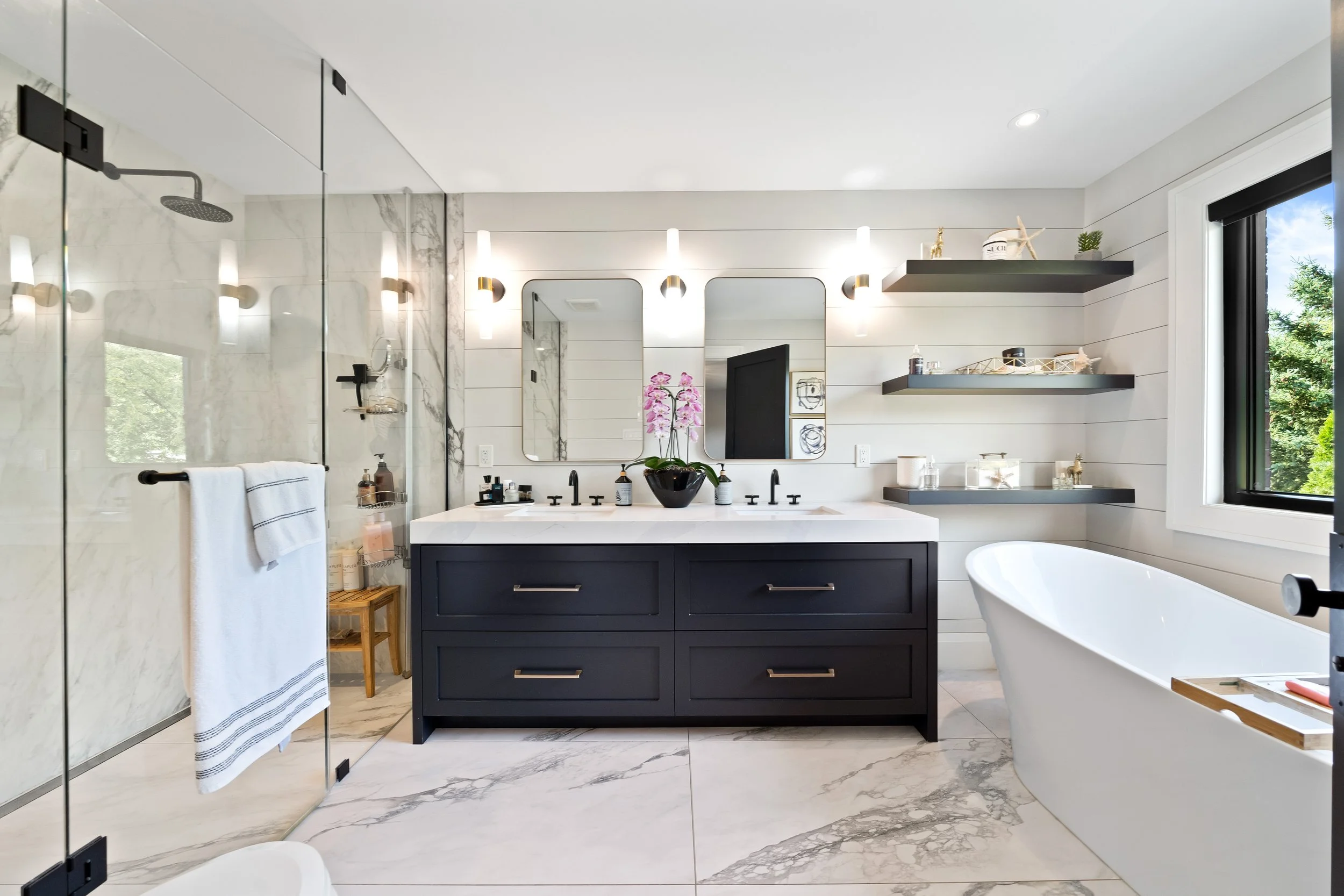 Modern bathroom with double vanity, large mirrors, black fixtures, white countertops, glass shower, free-standing bathtub, and open shelves with decor, natural light from window.
