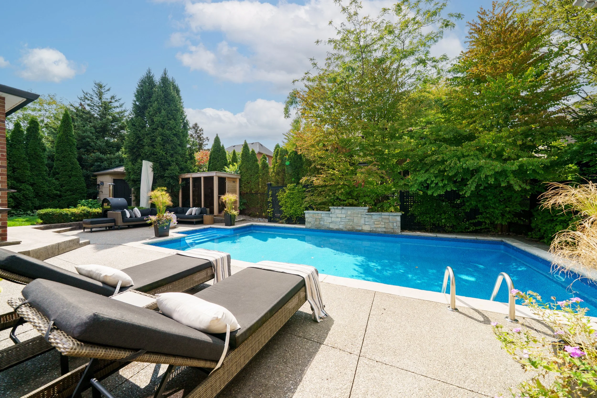 Backyard with a swimming pool, lounge chairs, a cabana with seating, lush green trees, and a partly cloudy sky.