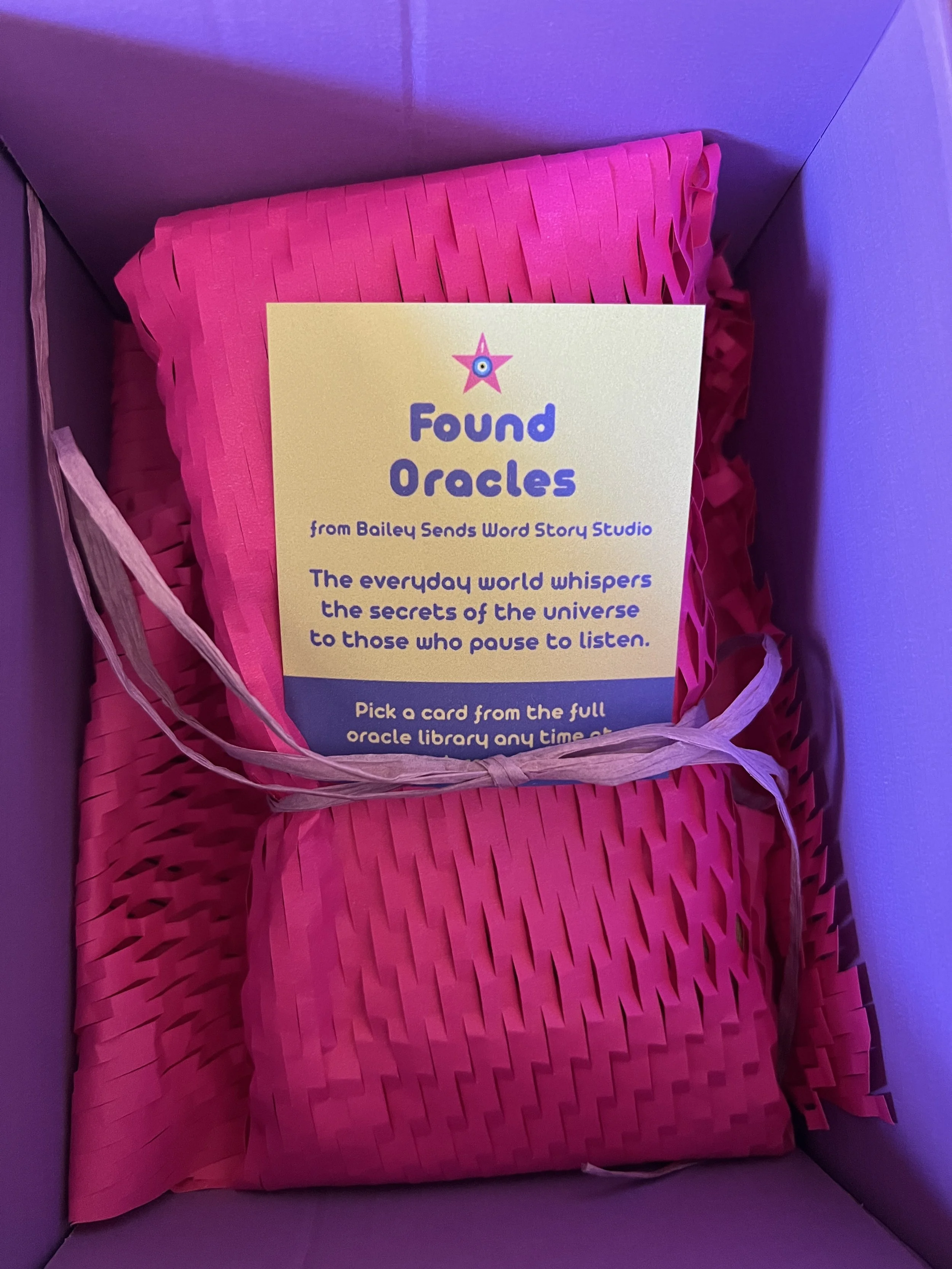 The wrapped Hello World mini deck from Found Oracles, tied up in hot pink honeycomb paper with a purple raffia bow and a card introducing the Found Oracles project and the deck on top.