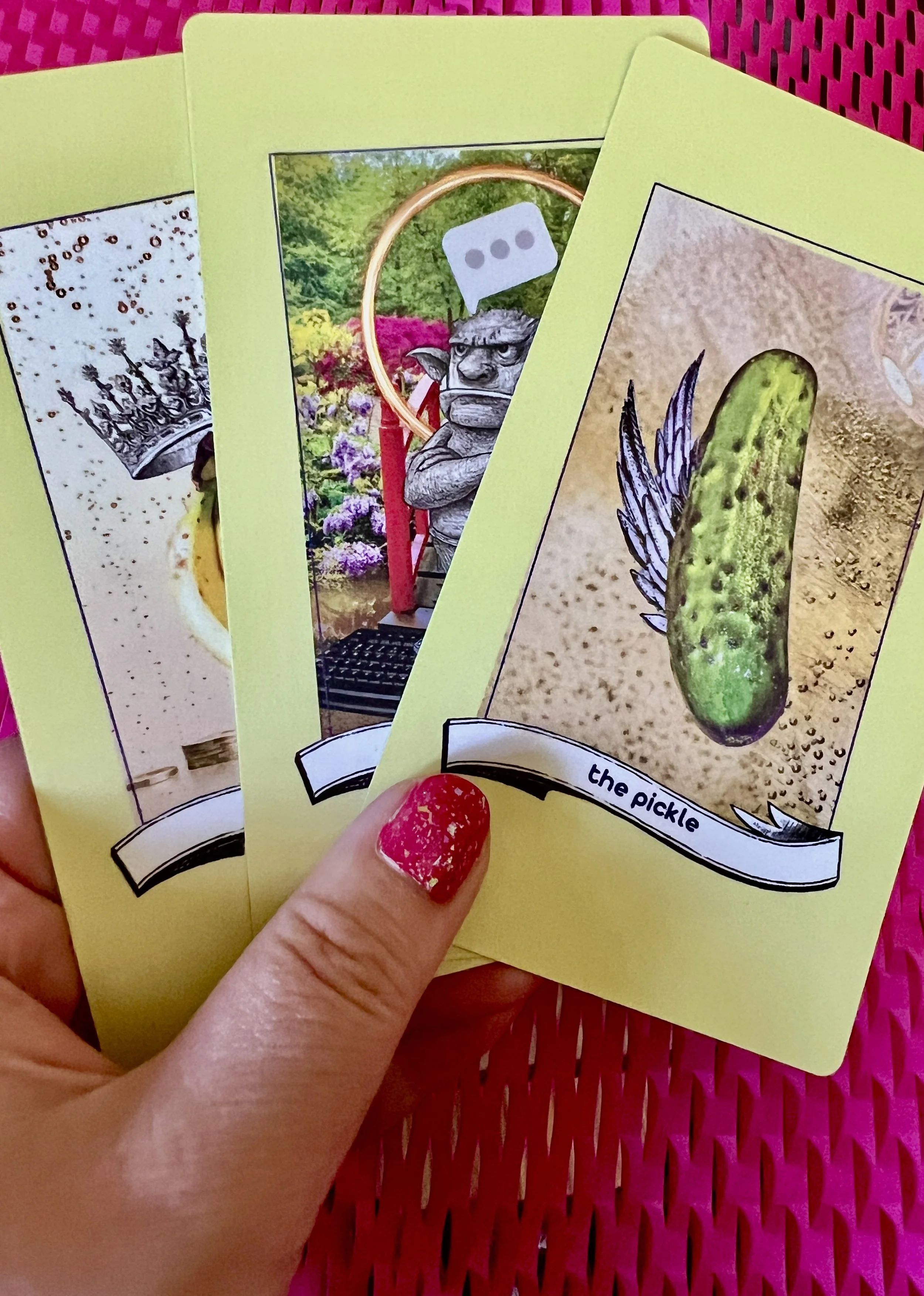 The artist's hand holding three of the cards from the Found Oracles Hello World mini deck - The Bananas, Teh Troll, and The Pickle on a hot pink honeycomb paper background.