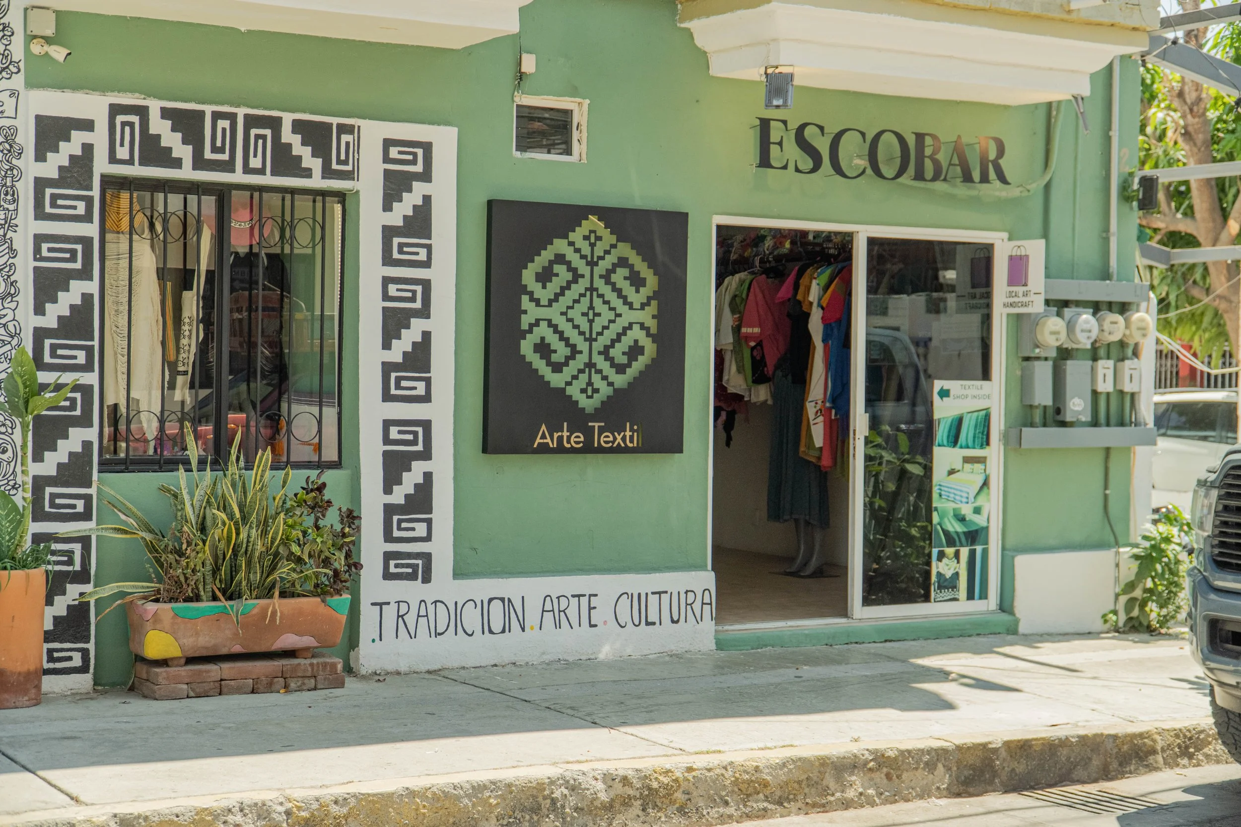 Fachada de tienda con cartel que dice 'Escobar' y otra que dice 'Arte Texti'. Hay plantas en macetas y una puerta de entrada con ropa en exhibición. Escritura en la pared que dice 'Tradición. Arte. Cultura'.