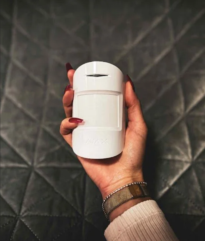 A hand holding a small white device with a slot at the top, against a gray tiled background.