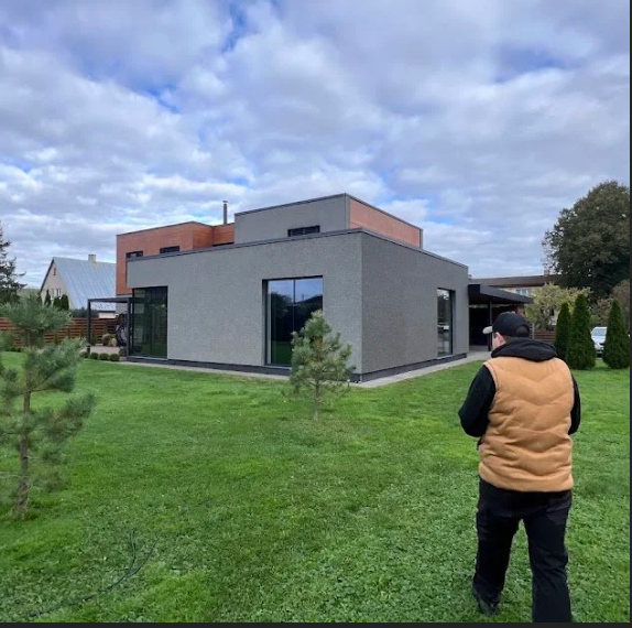 A person in a black hoodie and tan vest standing on a grassy area near a modern house with gray and orange exterior walls, large windows, and a cloudy sky overhead.