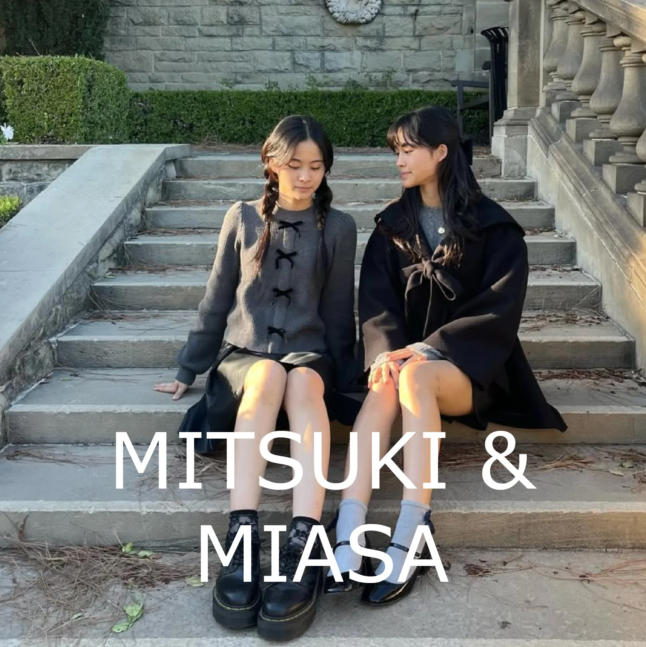 Two young women sitting on outdoor stone steps, one with braided hair wearing a grey sweater with black bows and a black skirt, the other with long dark hair wearing a black coat and grey socks with black shoes. The background features greenery and a stone wall, and the text 'MITSUKI & MIASA' is overlaid at the bottom.