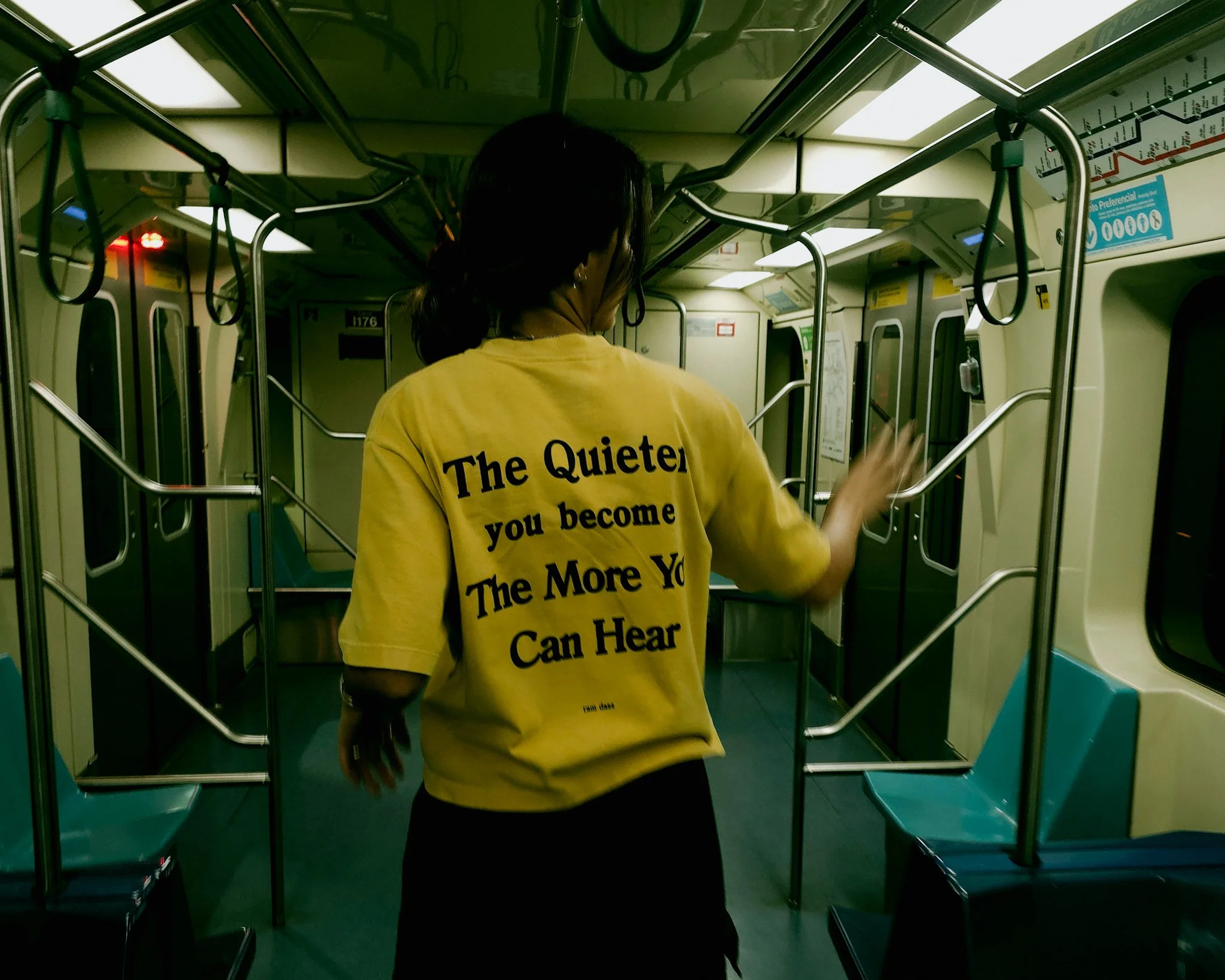 Pessoa de cabelo escuro vestindo uma camiseta amarela dentro de um transporte público, com o verso da camiseta visível, que tem a mensagem: "The Quieter you become The More You Can Hear"