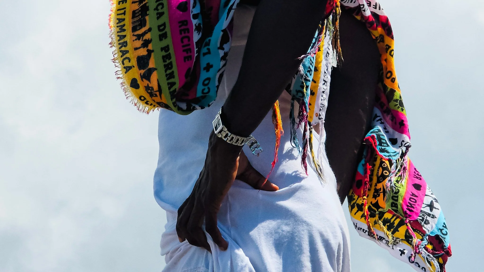 Pessoa com roupas coloridas segurando uma manta com padrões e palavras, na rua, de costas.