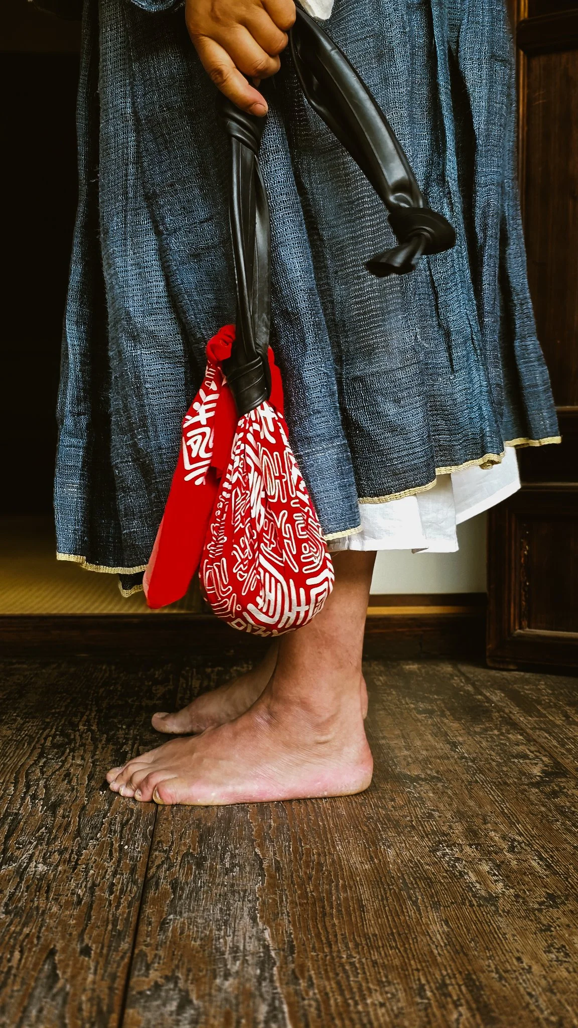 Pessoa de pé descalça, com uma saia azul escura, segurando um guarda-chuva vermelho com desenho branco, dentro de uma casa com piso de madeira.