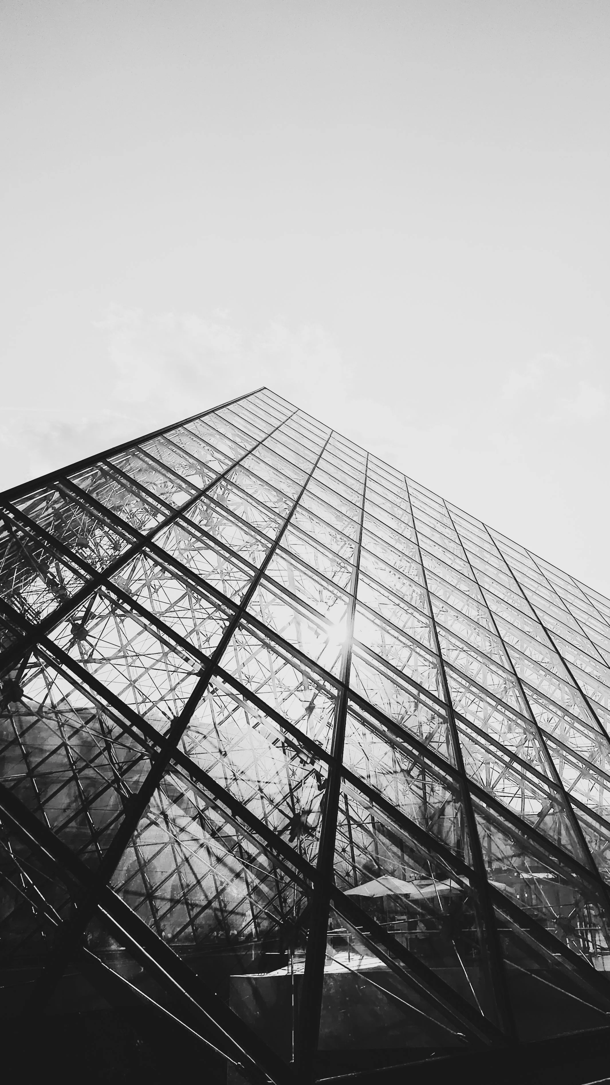 Imagem em preto e branco de um edifício de estrutura de vidro, visto de baixo para cima, com o sol refletido nas janelas.