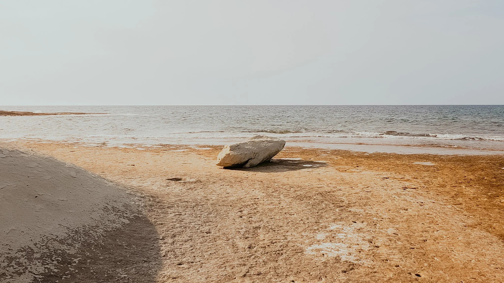 Praia com areia marrom, uma grande pedra na areia e o mar ao fundo com ondas suaves, céu claro, sem pessoas.