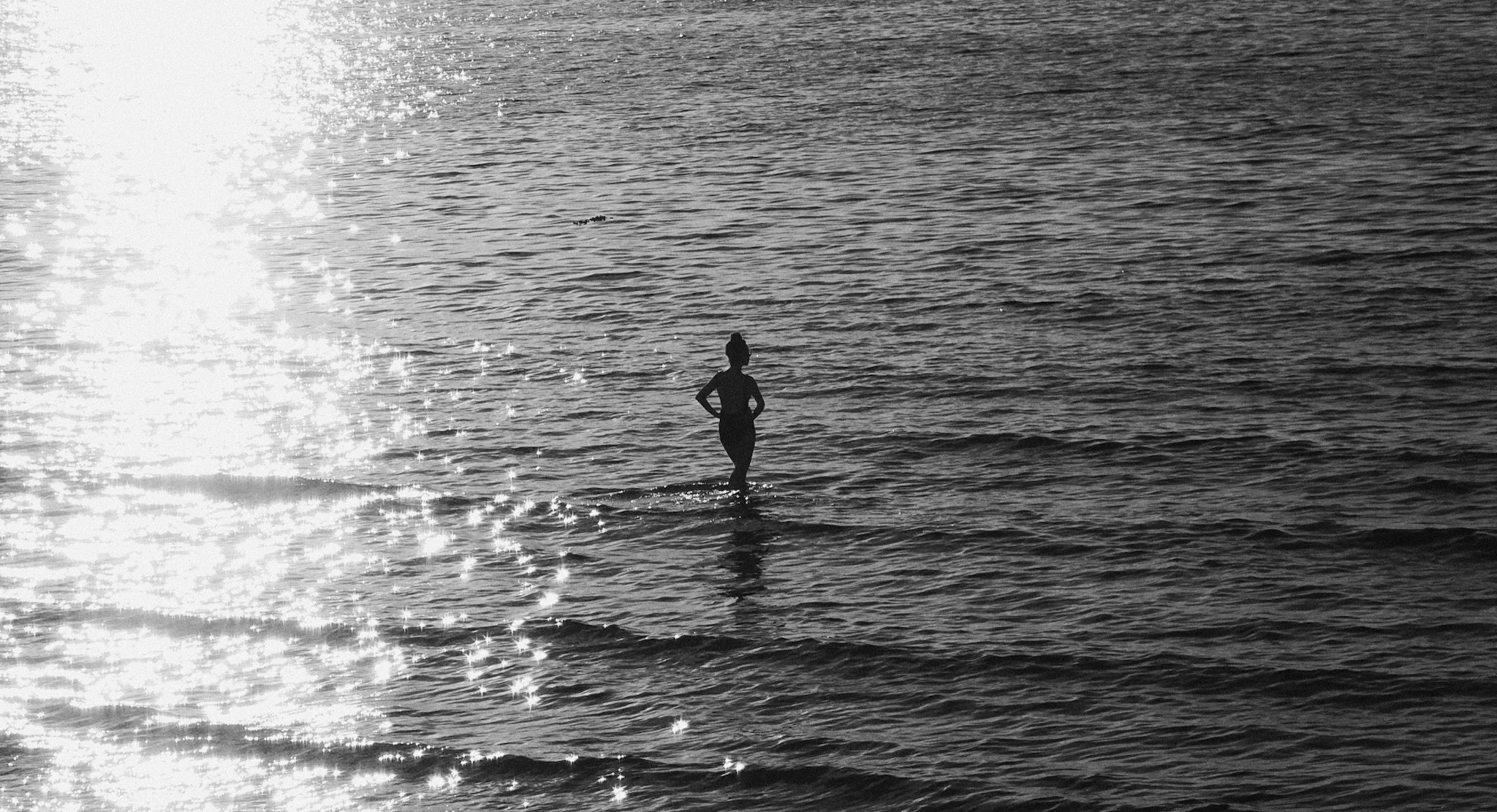 Silhueta de uma mulher em pé na água do mar com reflexos brilhantes ao sol.