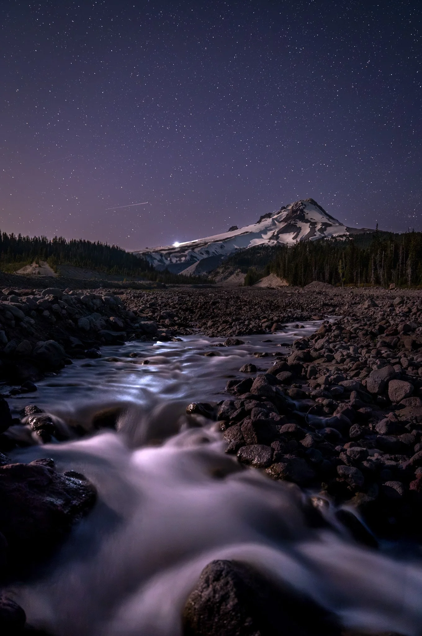 Nighttime landscape of a mountain with snow, a starry sky with visible shooting stars, a flowing river with smoothwater effect, and a forested area.
