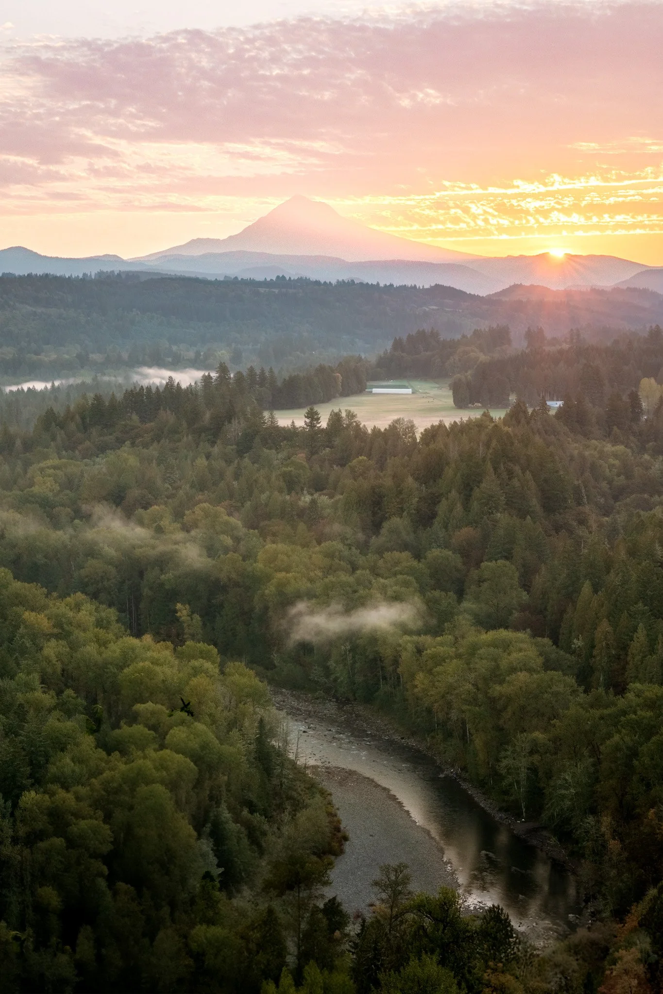 Sunrise over a mountainous landscape with lush green trees, a river, and a distant volcano in the background.