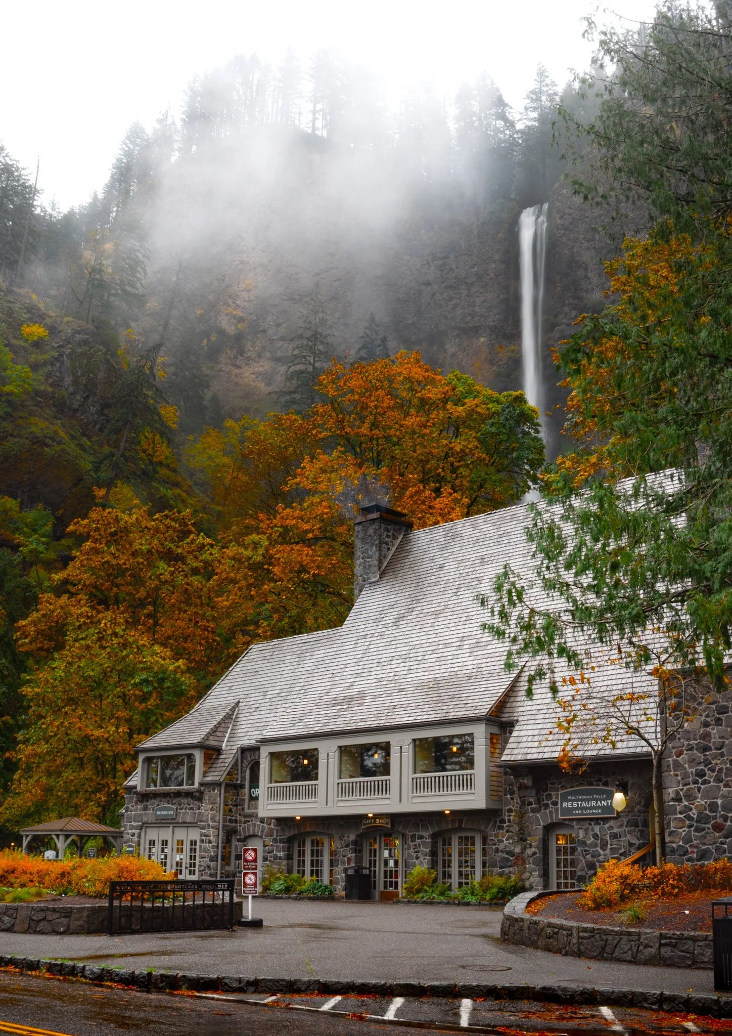 A stone building with large windows and a balcony, surrounded by fall foliage, with Multnomah Falls waterfall cascading in the background among misty trees.