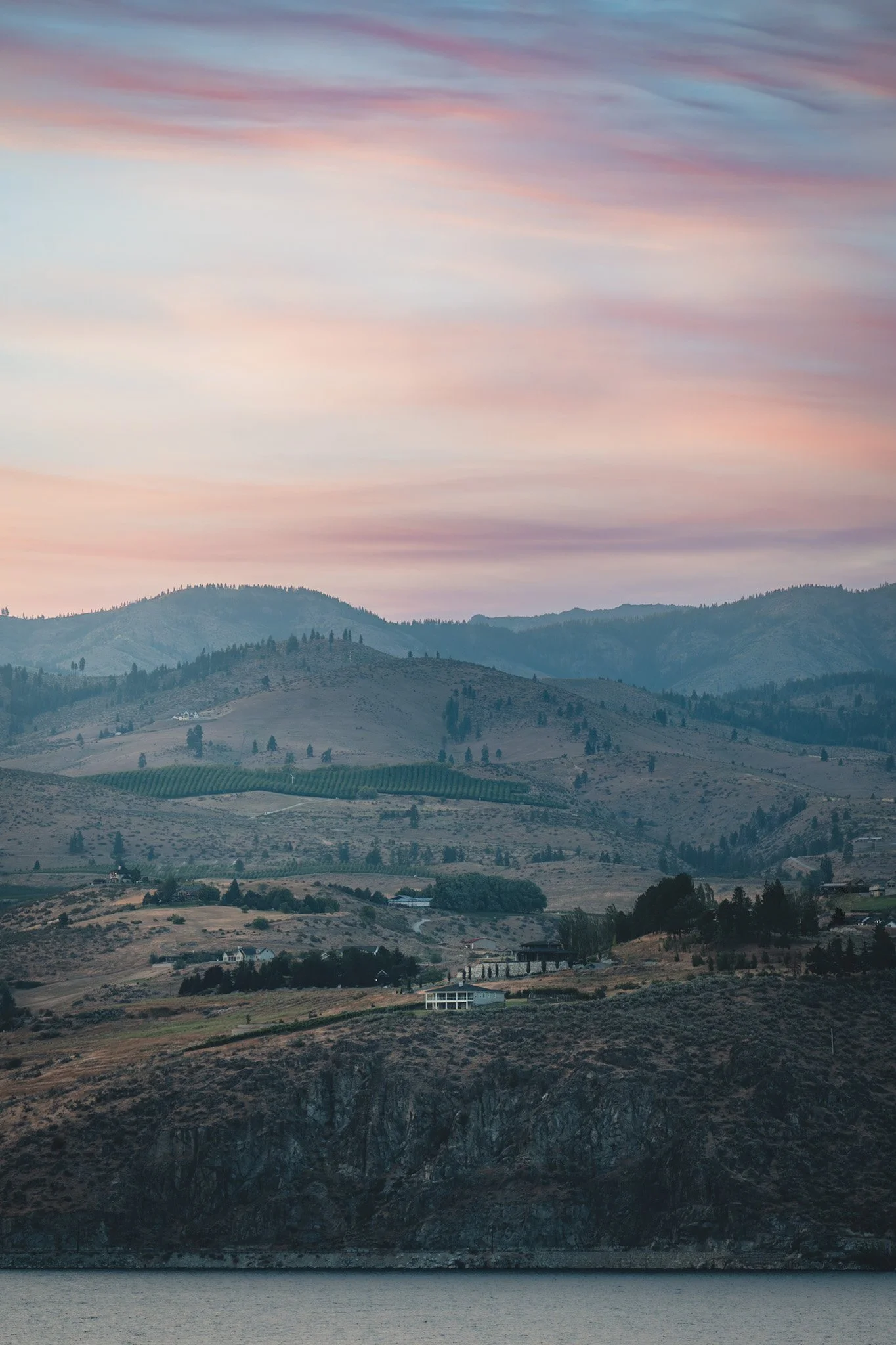 A scenic view of rolling hills and mountains at dusk with a colorful sky, some houses, and a body of water in the foreground.