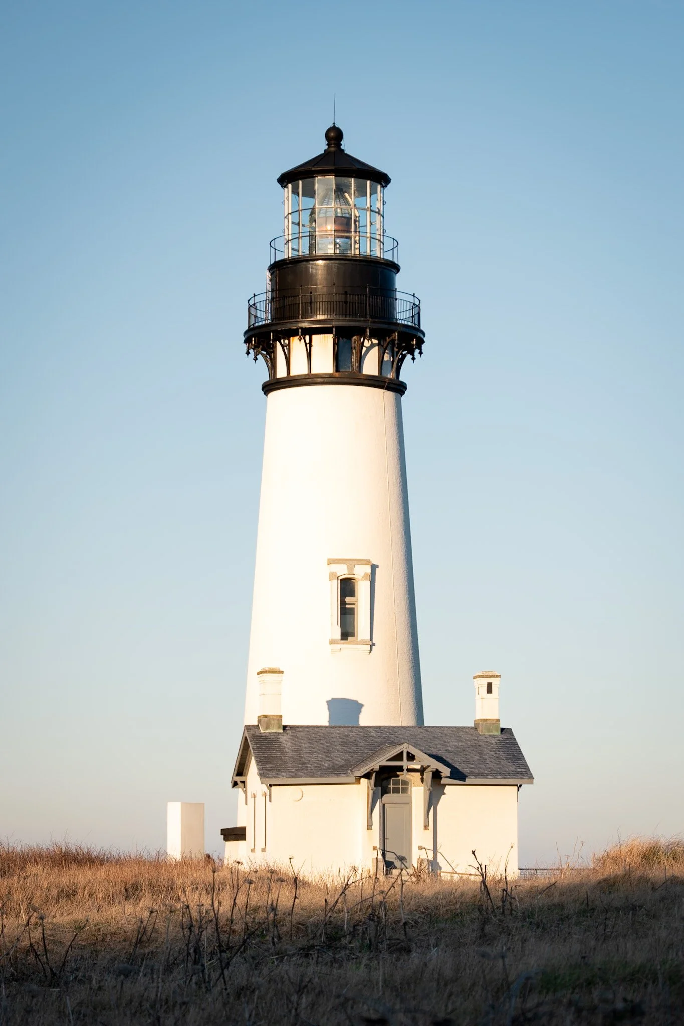 A tall lighthouse with a black top and railing, white body, and small adjacent building with a door, set in a grassy area under a clear blue sky.