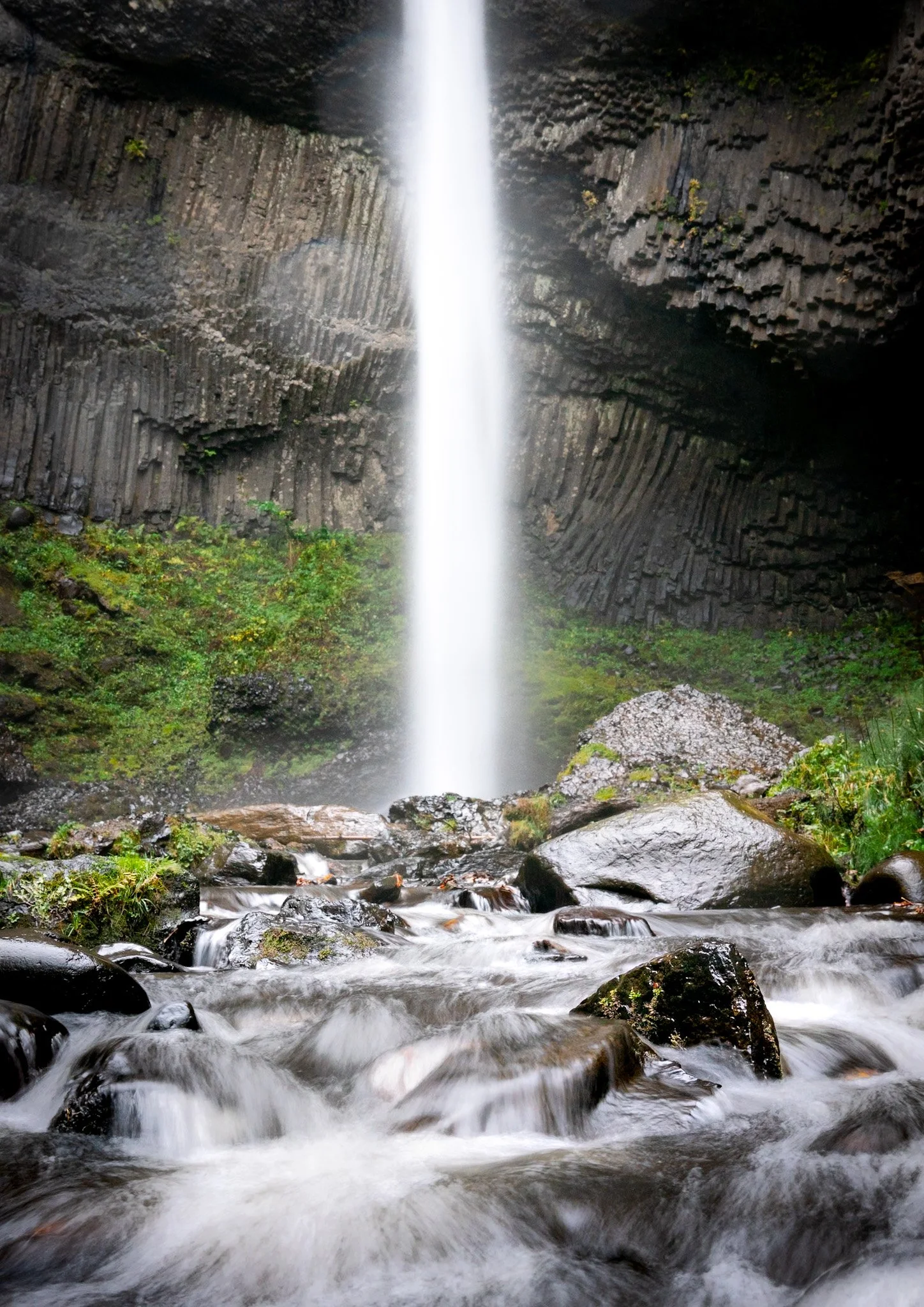 A tall, narrow waterfall flowing from a cliff into a rocky stream below amidst greenery.