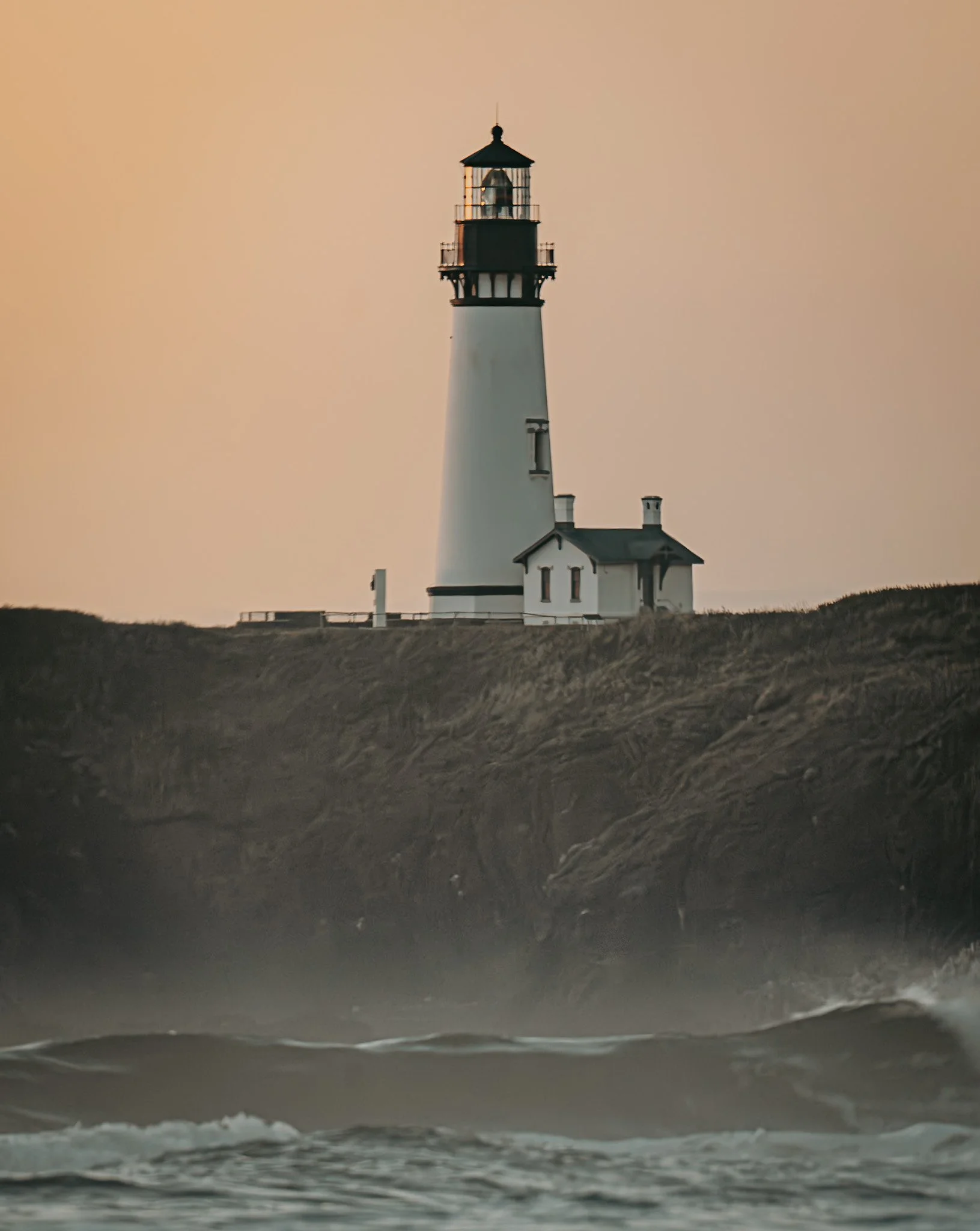 Lighthouse on a rocky coast at sunset with waves crashing below.
