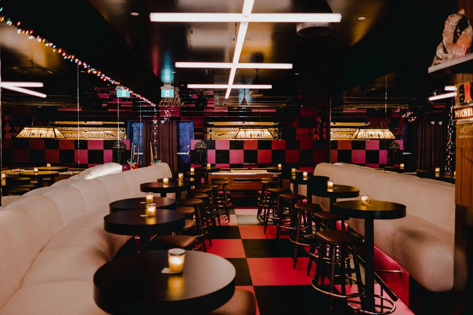 Intérieur d'un restaurant ou bar avec des tables rondes, des chaises, des banquettes blanches, des bougies allumées sur chaque table, décorations lumineuses et mur aux motifs rouges et noirs.