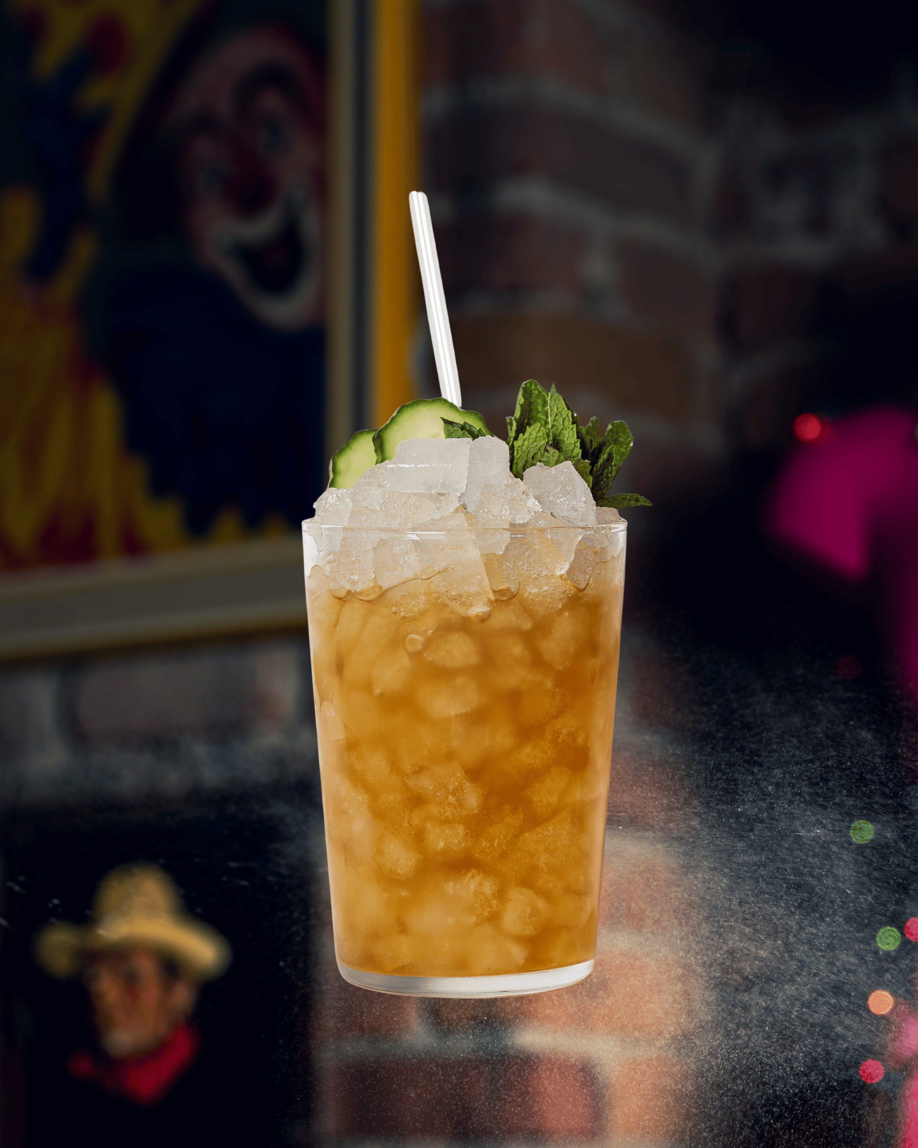 Un verre de cocktail glacé avec des tranches de concombre et une branche de menthe en garniture, avec une paille blanche, posé sur un comptoir en surface sombre, avec une peinture de clown en arrière-plan.