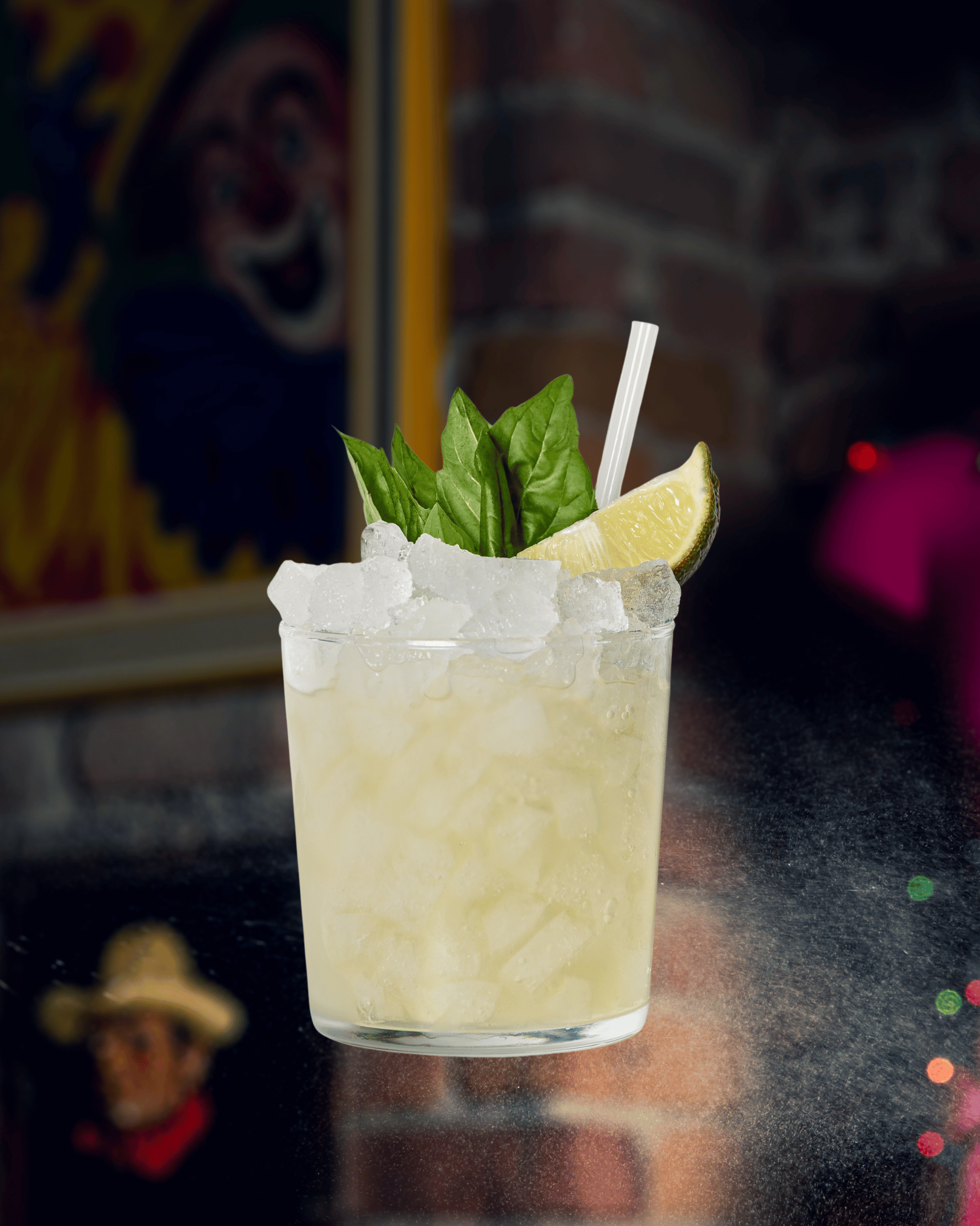 Un verre de cocktail gelé avec du citron, des feuilles de basilic et une paille, posé sur une surface sombre avec un décor de fête en arrière-plan.