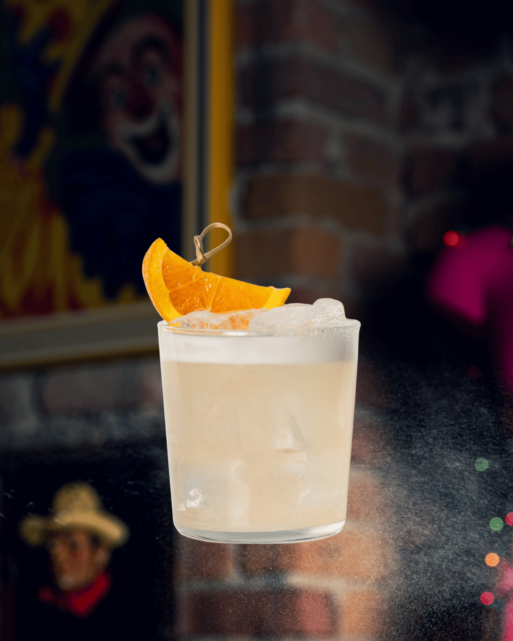 Cocktail clair avec glaçons, garniture d'une tranche d'orange et un petit cure-dent, sur un fond flou avec un portrait de clown et des murs en briques
