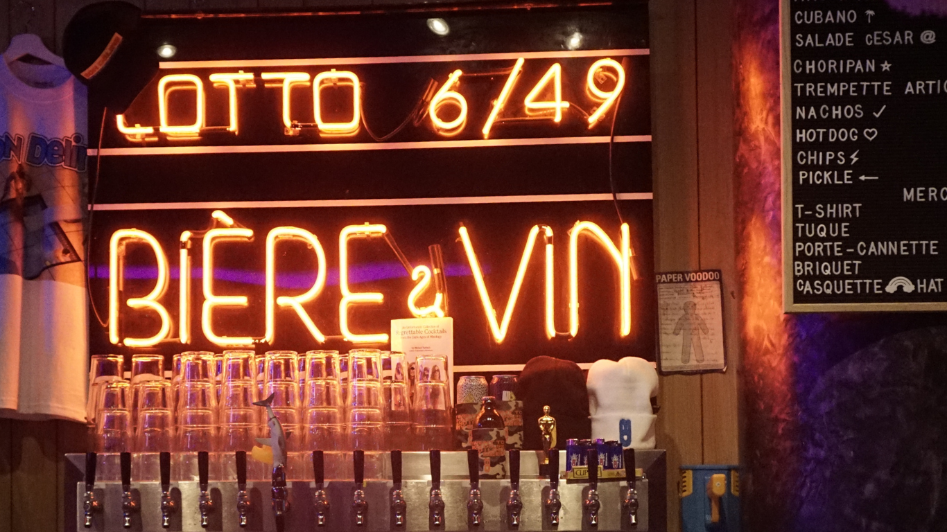Un panneau lumineux en néon orange affiche le prix de la bière à 6,49 euros. En dessous, il y a des verres empilés et une rangée de robinets de bière. À droite, un menu écrit à la craie indique différents plats et boissons.