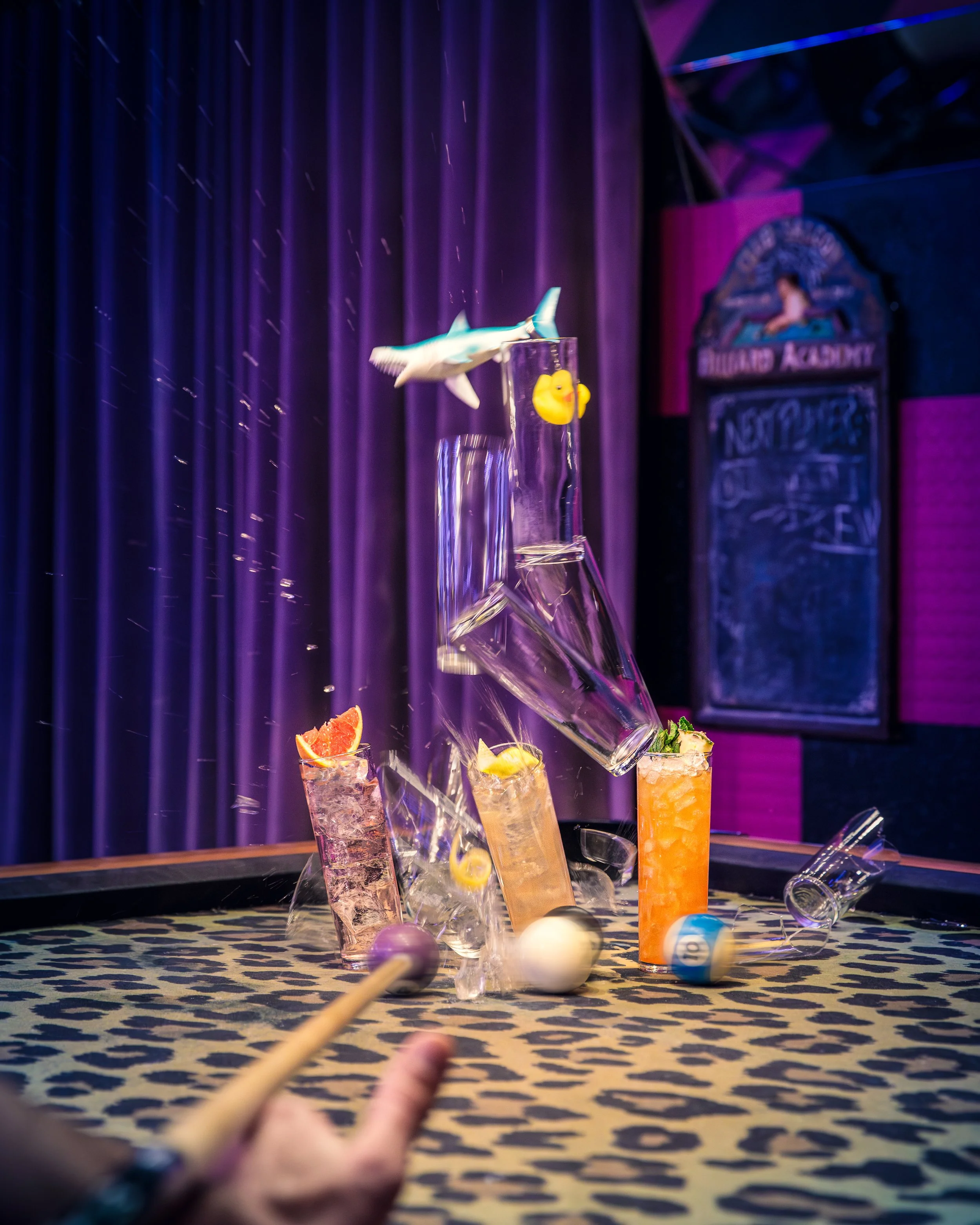 Cliché d'une scène de jonglage avec des verres de cocktails, des balles de billard, et un jouet de poisson, au plafond d'un spectacle ou bar, avec un fond de rideau violet et un tableau noir