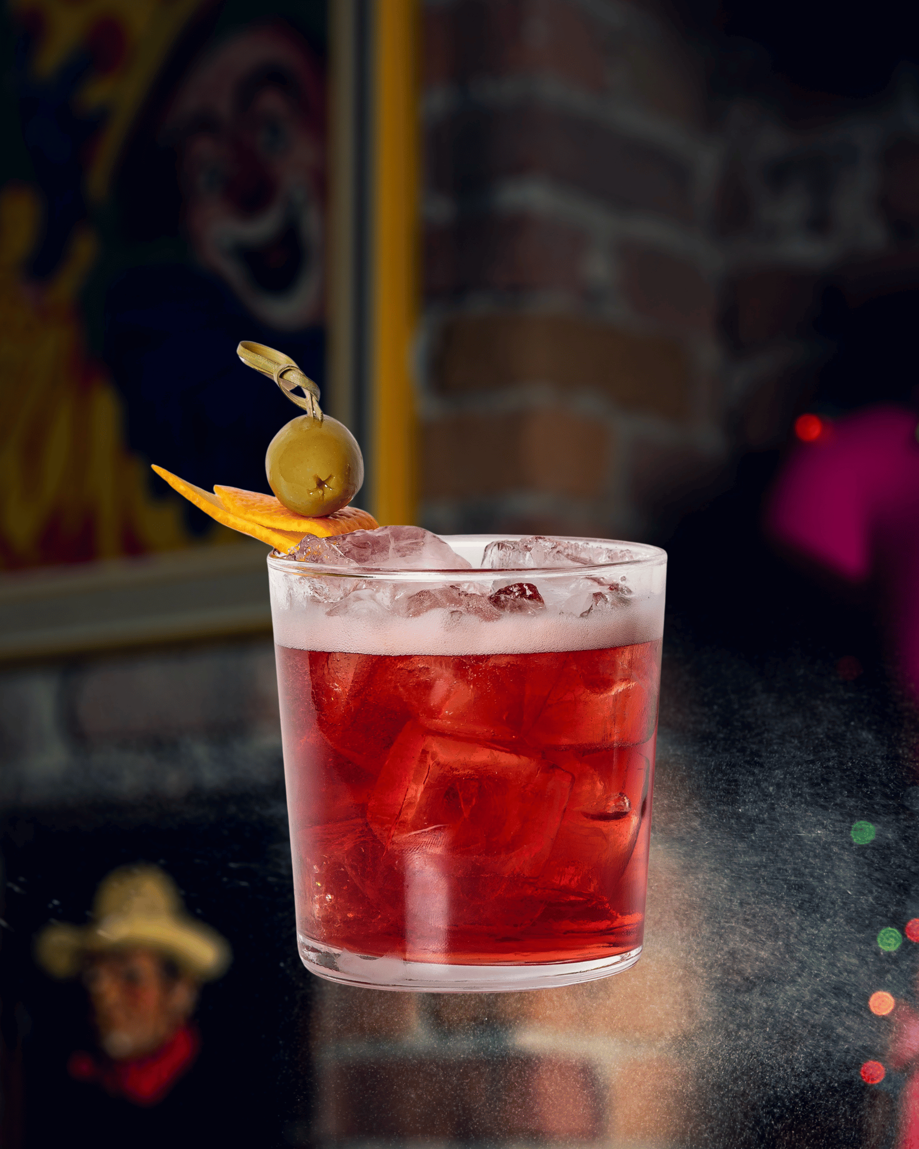 Un verre de cocktail rouge avec glaçons, décoré d'une olive verte sur un cure-dent et d'une tranche d'orange, sur un fond sombre et flou.