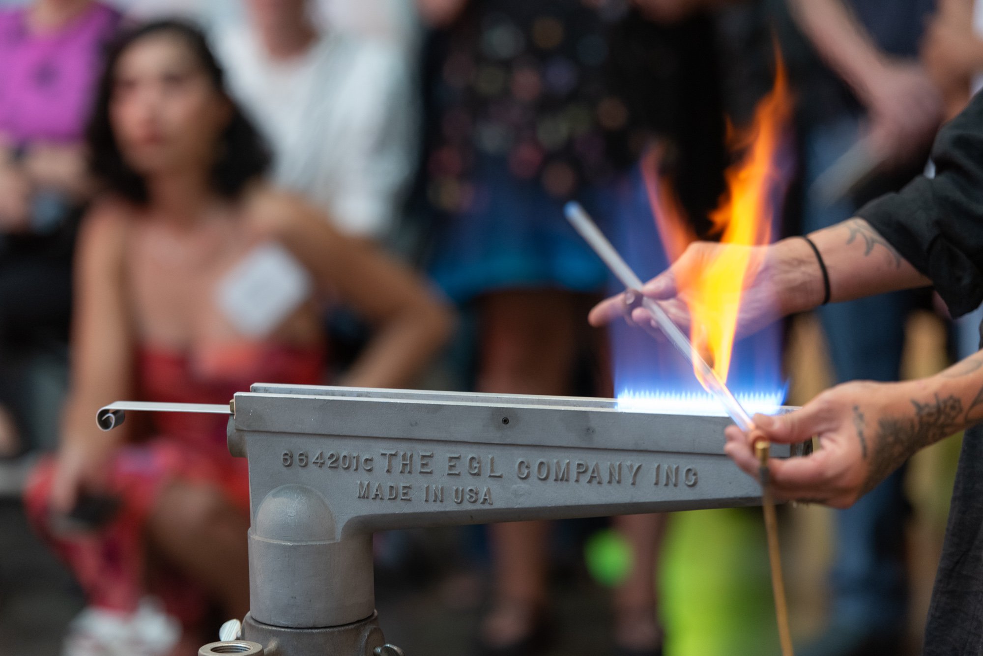 Person holding a glass tube in a ribbon burner flame, surrounded by onlookers at a demonstration event.