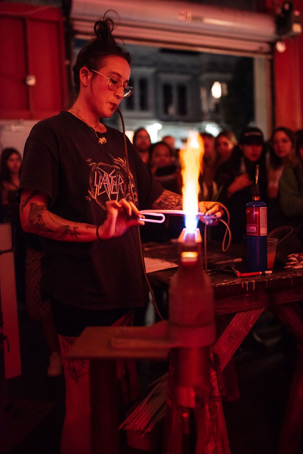 A woman with glasses and tattoos creating a flame with a flame-throwing tool in a dimly lit room filled with people.