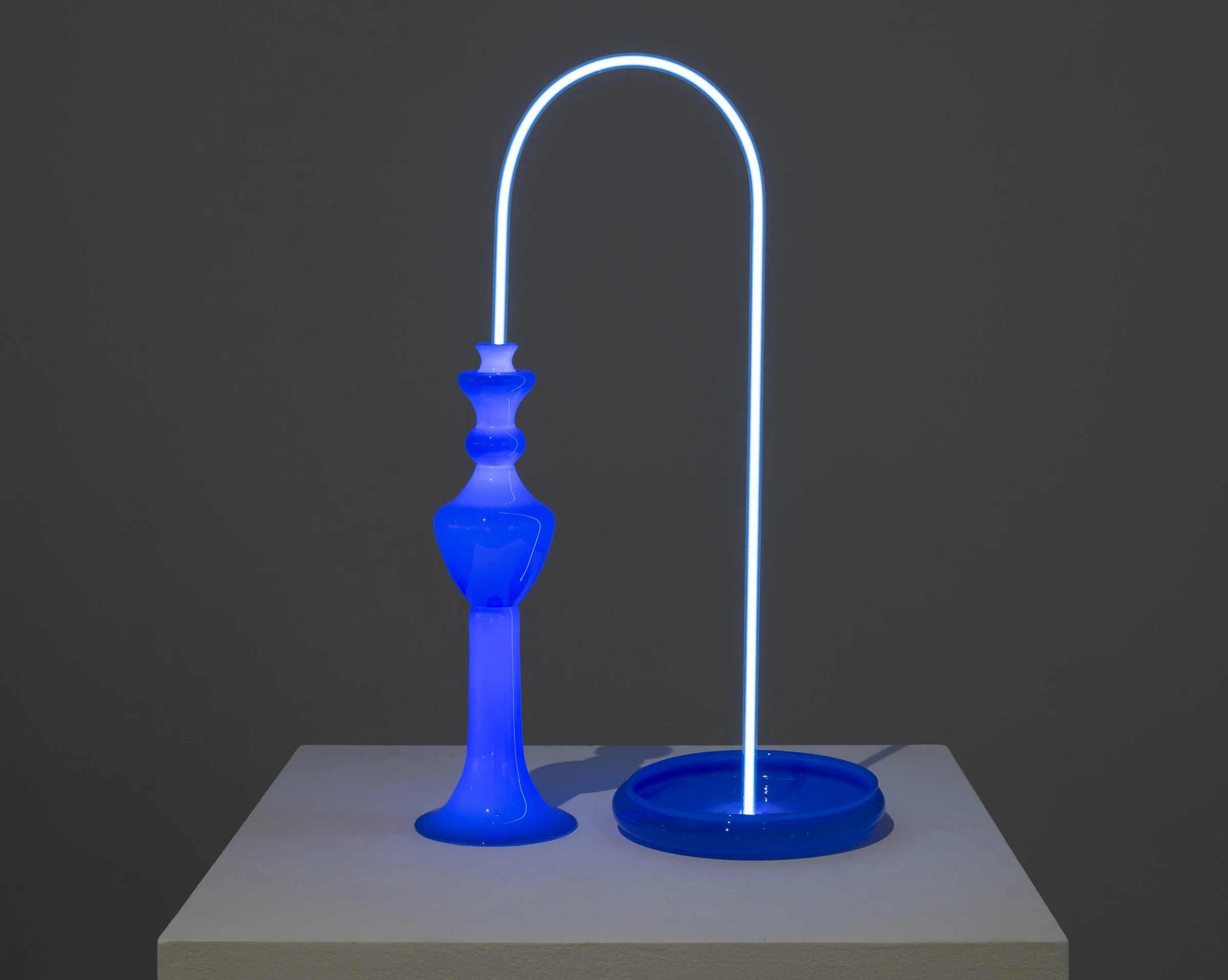 Blue glass sculpture resembling a candlestick and a tall illuminated blue glass tube art piece on a white pedestal.