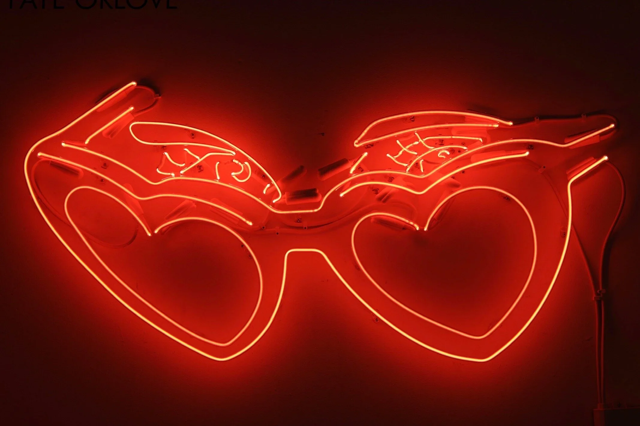Neon sign of two sunglasses with heart-shaped lenses, glowing red-orange.