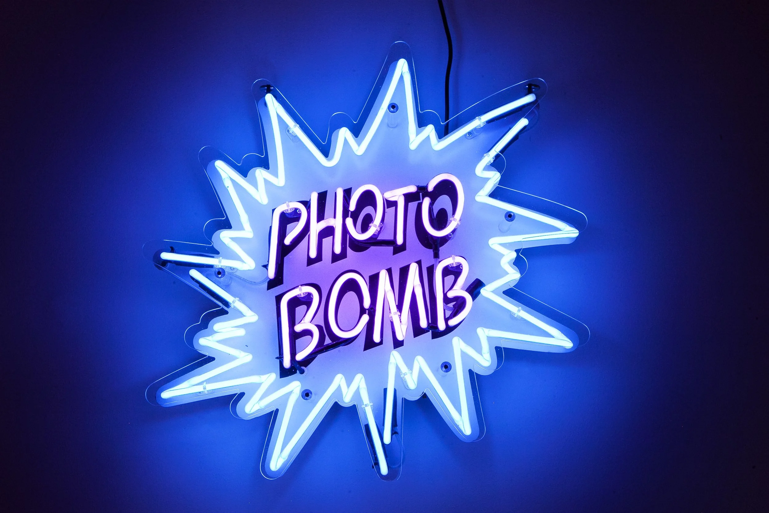 Neon sign with purposefully reversed text reading 'PHOTO BOM' inside a starburst shape, glowing in blue and purple hues.