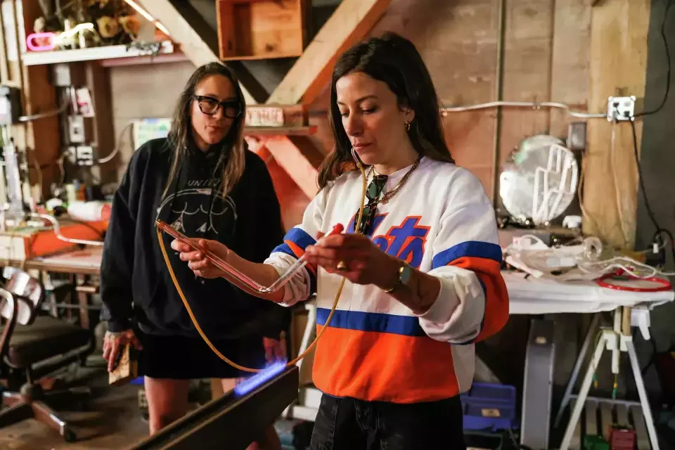 Two women in a neon studio, one is holding a glass tube connected to a blow hose, the other watches. Shelves and tools are visible in the background.