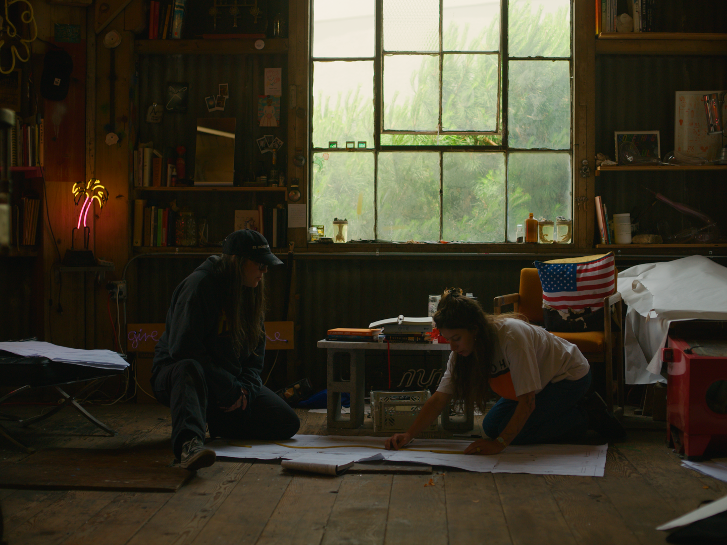 Two women working on large papers or plans on the wooden floor of a rustic room with a large window, bookshelves, and various objects on shelves. One woman is kneeling and the other is crouching, both focused on the plans. The room has natural light, a neon palm tree sign, and an American flag draped over a chair.